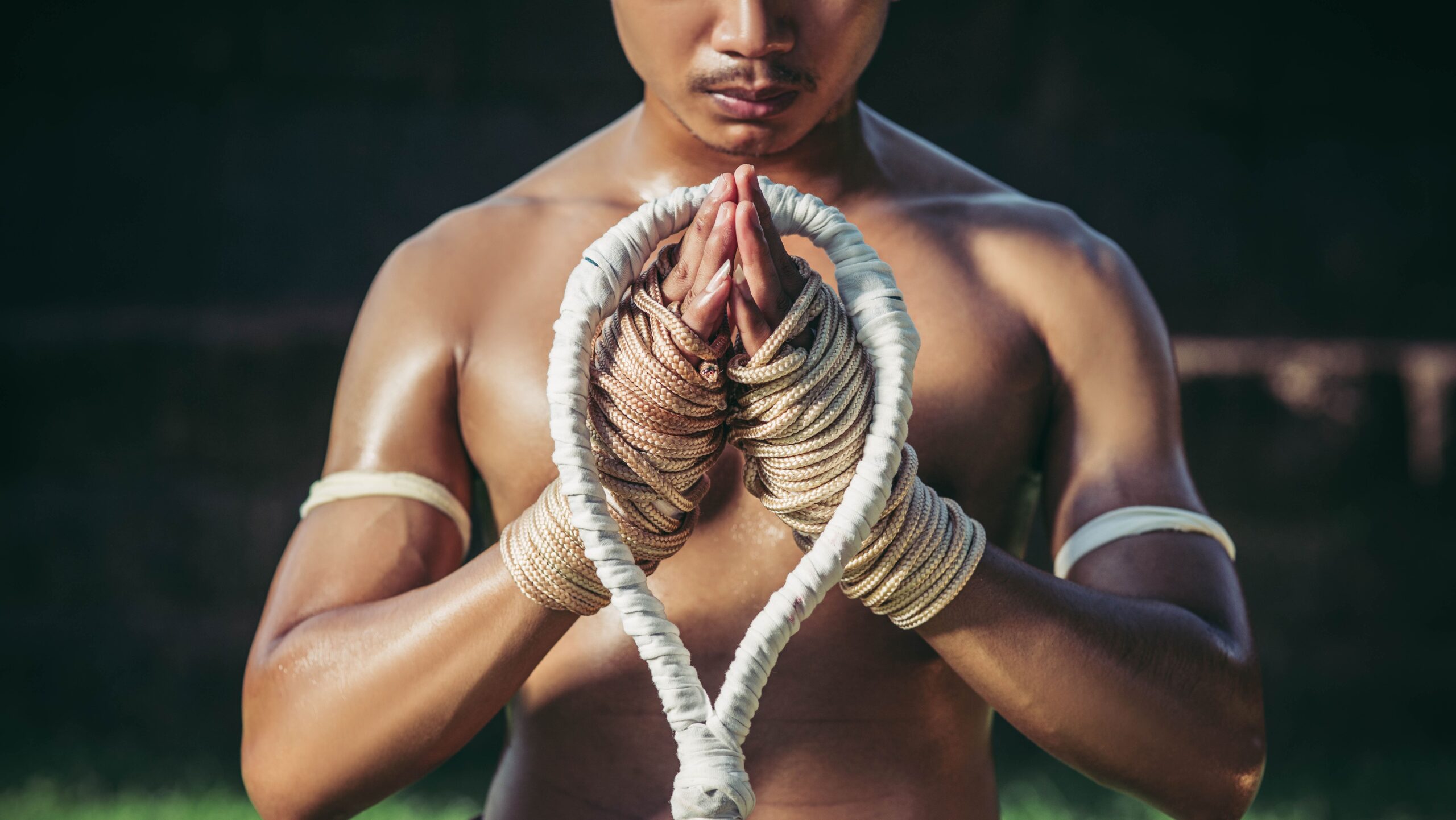 Praticante de Muay Thai com as mãos envoltas em cordas beges unidas em cumprimento tradicional, segurando um mongkhon trançado à frente do peito.