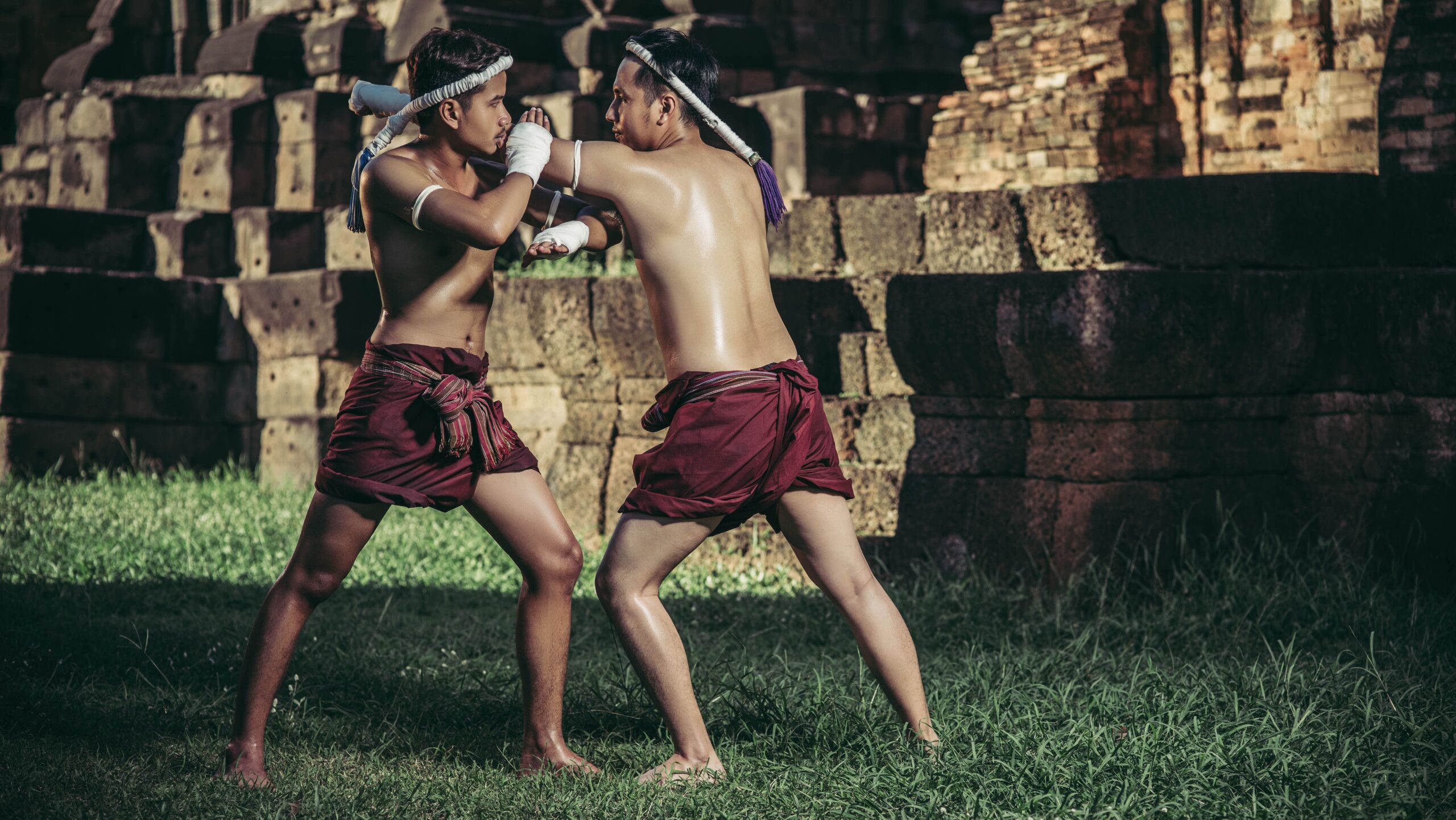 Dois lutadores de muay thai tradicional em trajes históricos com mongkhon na cabeça, demonstrando a postura correta, base equilibrada e posicionamento dos braços ao ar livre, em um gramado diante de ruínas antigas de templo de pedra.