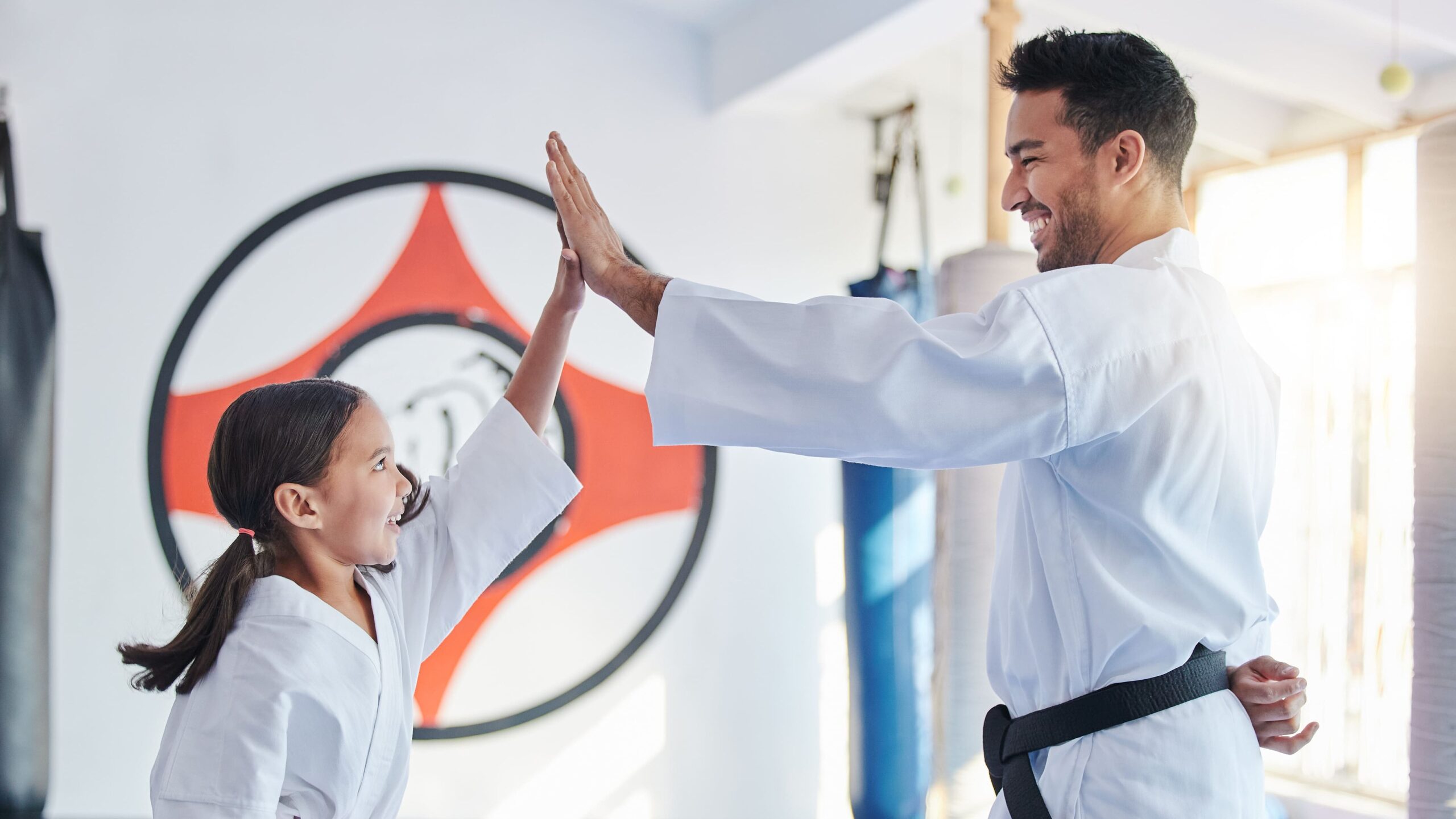 Aluna mirim de quimono com faixa laranja comemorando com um toque de mãos ("high five") com seu professor de faixa preta na academia.