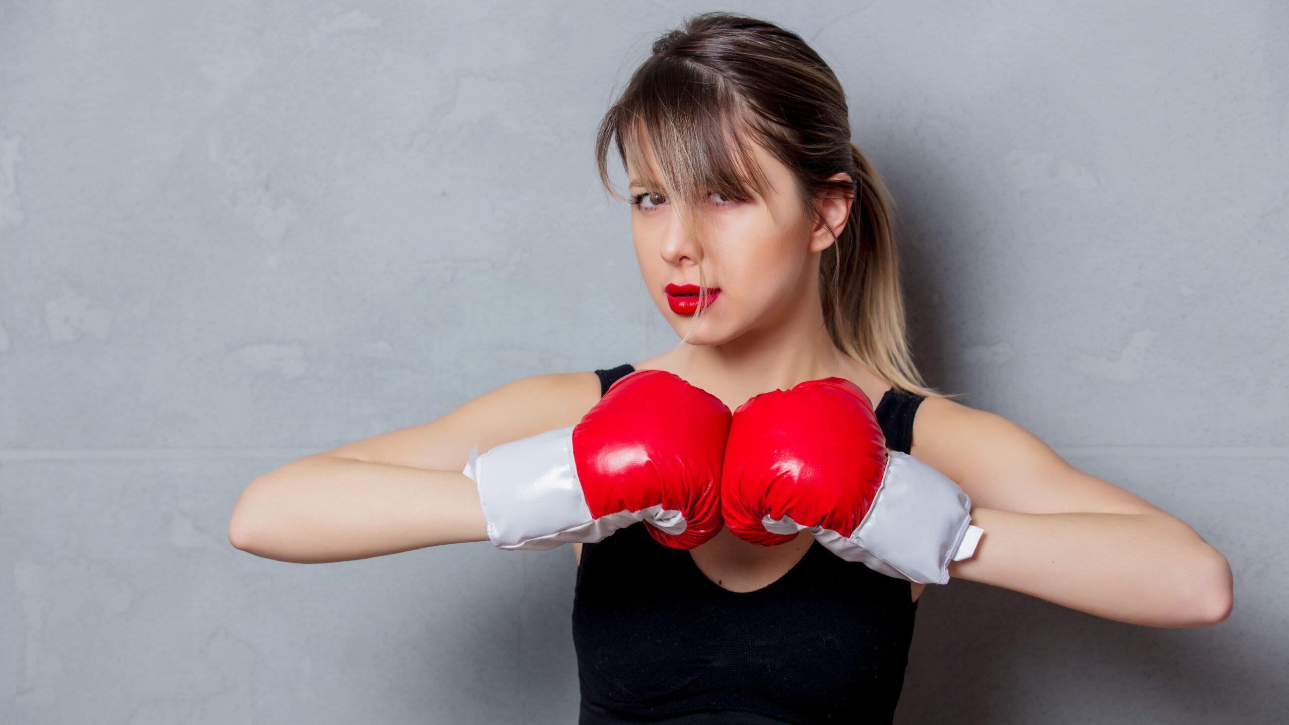 Mulher em estúdio, com cabelo preso e maquiagem marcante, usando luvas de boxe vermelhas e luvas brancas, em posição de combate com os braços à frente contra um fundo cinza.