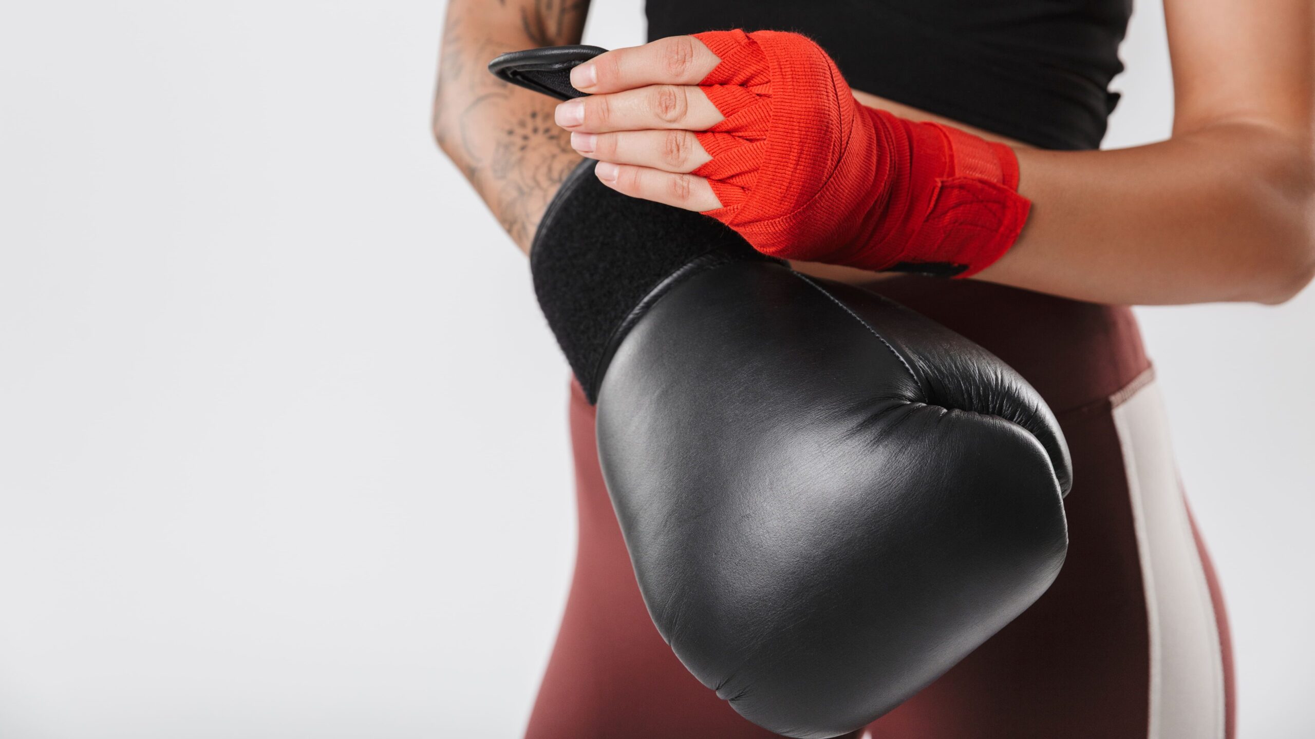 Lutador(a) com bandagem de pano vermelho no punho, usando luva de boxe preta, em pose de proteção junto ao corpo, com foco na mão enfaixada e na luva