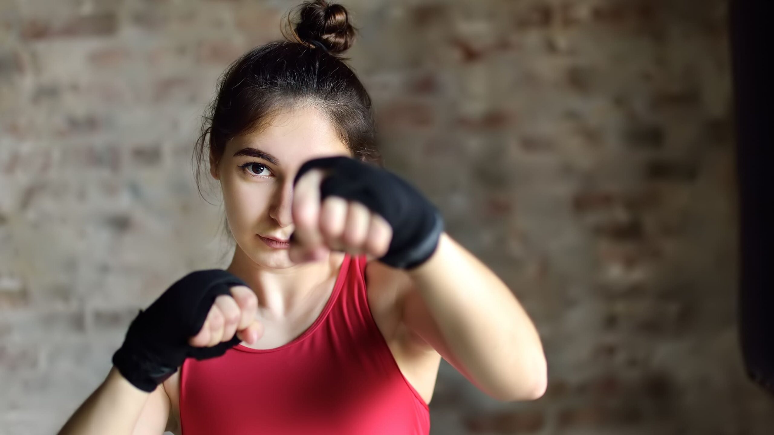 Mulher jovem em treino esportivo com top vermelho e faixa preta nos braços, fazendo exercício de empurrar com as mãos estendidas, em ambiente com parede de tijolos ao fundo.