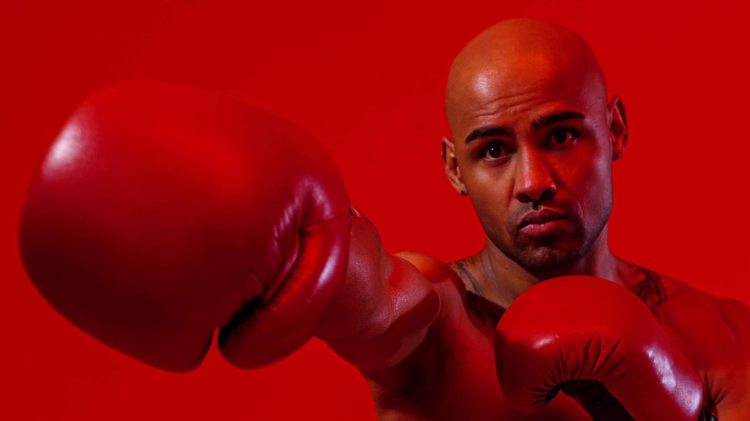 Homem com luvas de boxe vermelhas lançando um soco em direção à câmera em ambiente com iluminação vermelha.