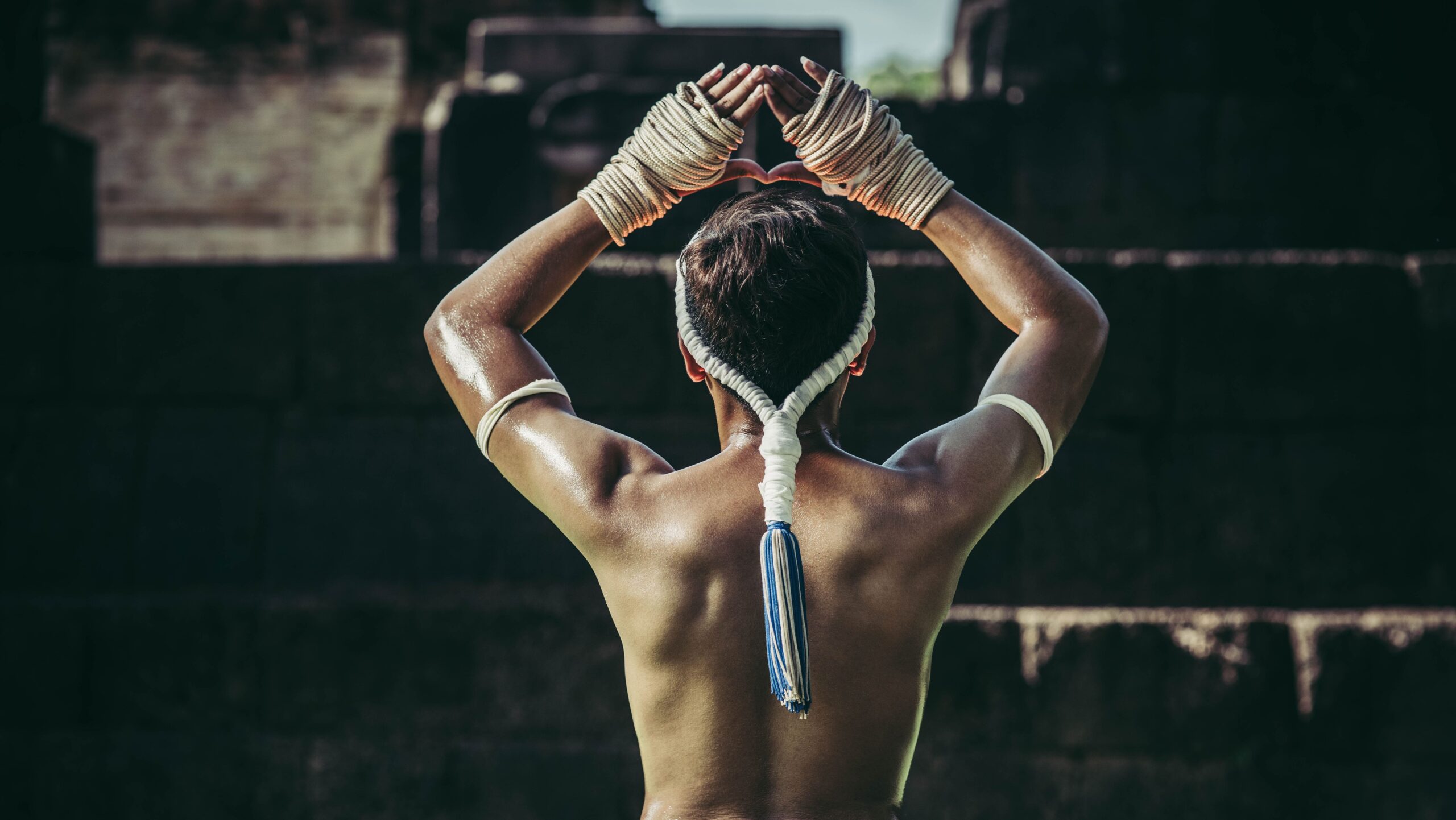 Lutador de Muay Thai visto de costas com as mãos erguidas, destacando o adorno cerimonial mongkhon com pendão azul e branco caindo pelas costas.