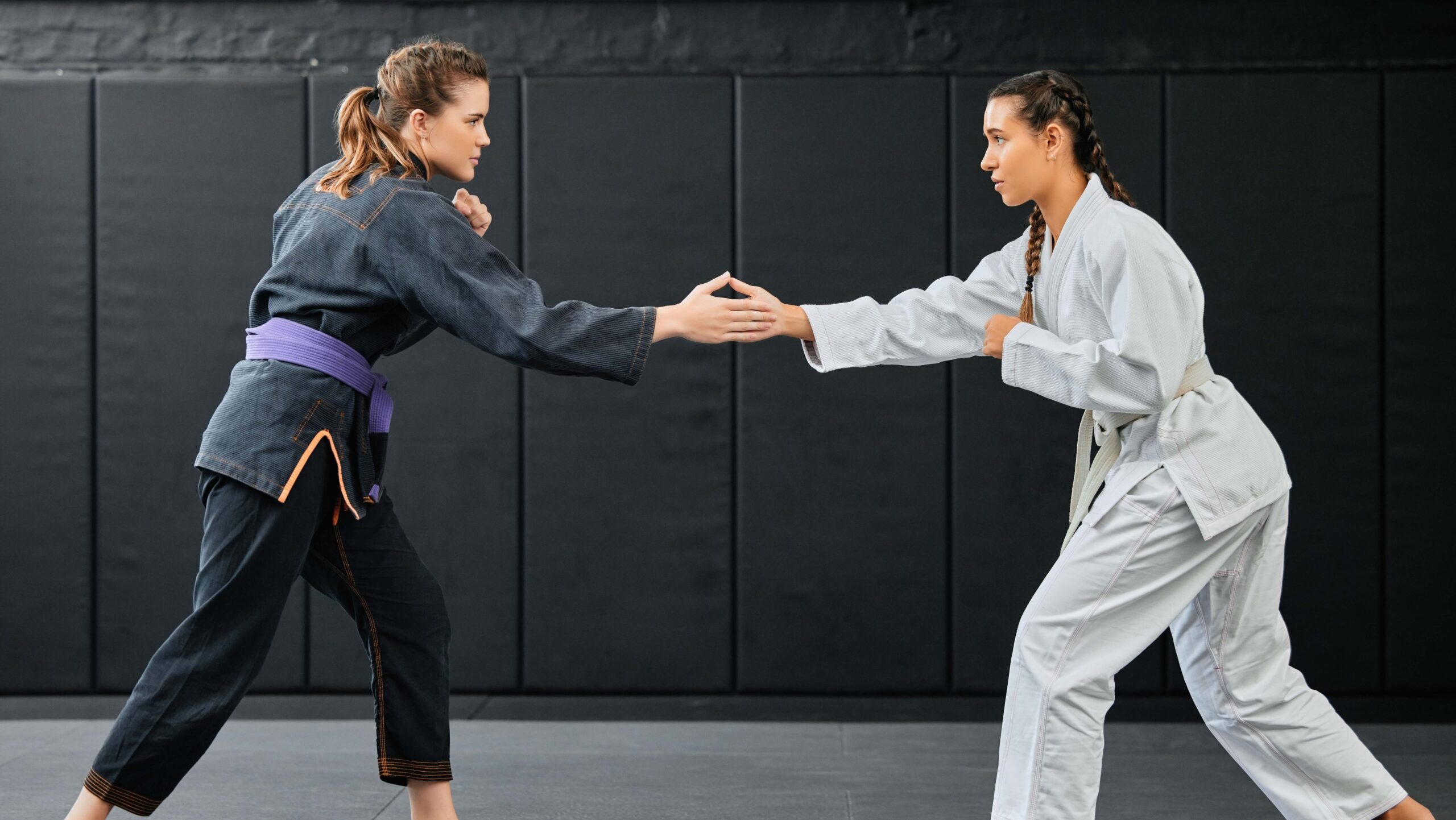 Duas mulheres treinando artes marciais em um tatame: uma de kimono escuro e outra de kimono branco, em posição de combate com as mãos estendidas, em ambiente externo com parede de fundo.