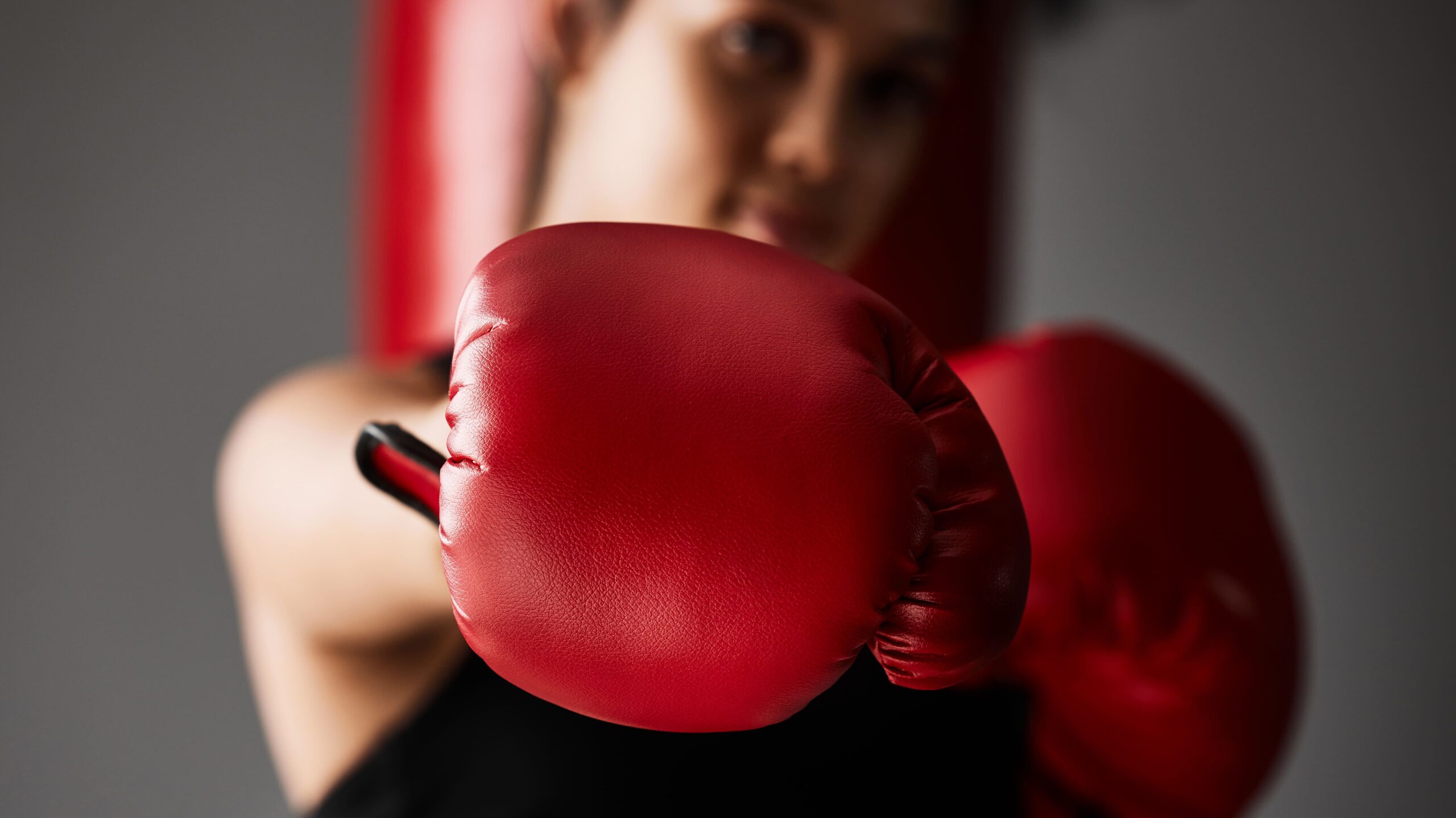 Homem com roupa de boxe em fundo desfocado, exibindo luvas de boxe vermelhas em primeiro plano, posição de treino e foco no combate esportivo.