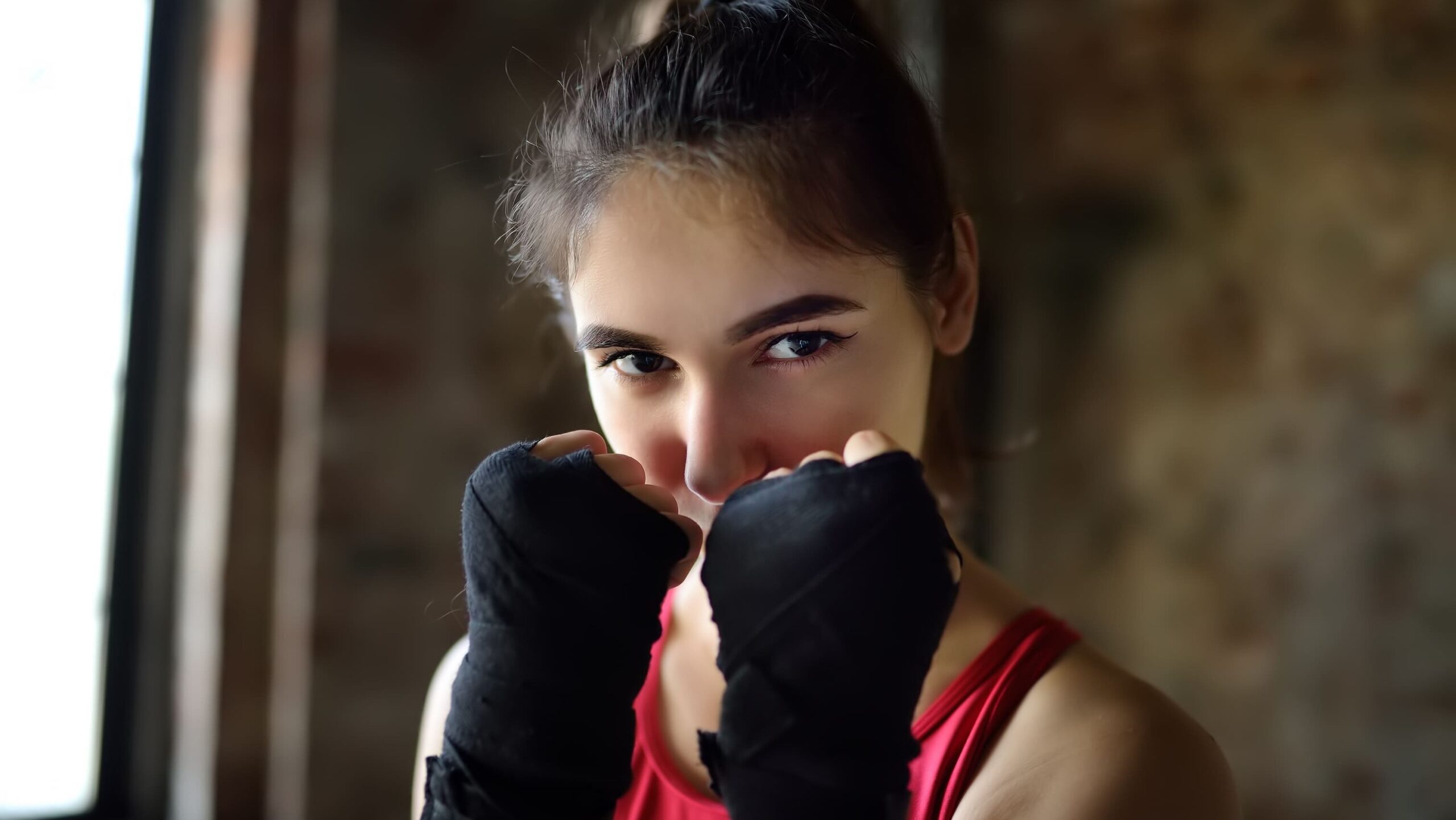 Retrato em iluminação suave de uma jovem praticante de muay thai feminino, com o cabelo preso, vestindo regata vermelha e com as mãos envolvidas em bandagens pretas levantadas na posição de guarda diante do rosto, olhando fixamente para a câmera.