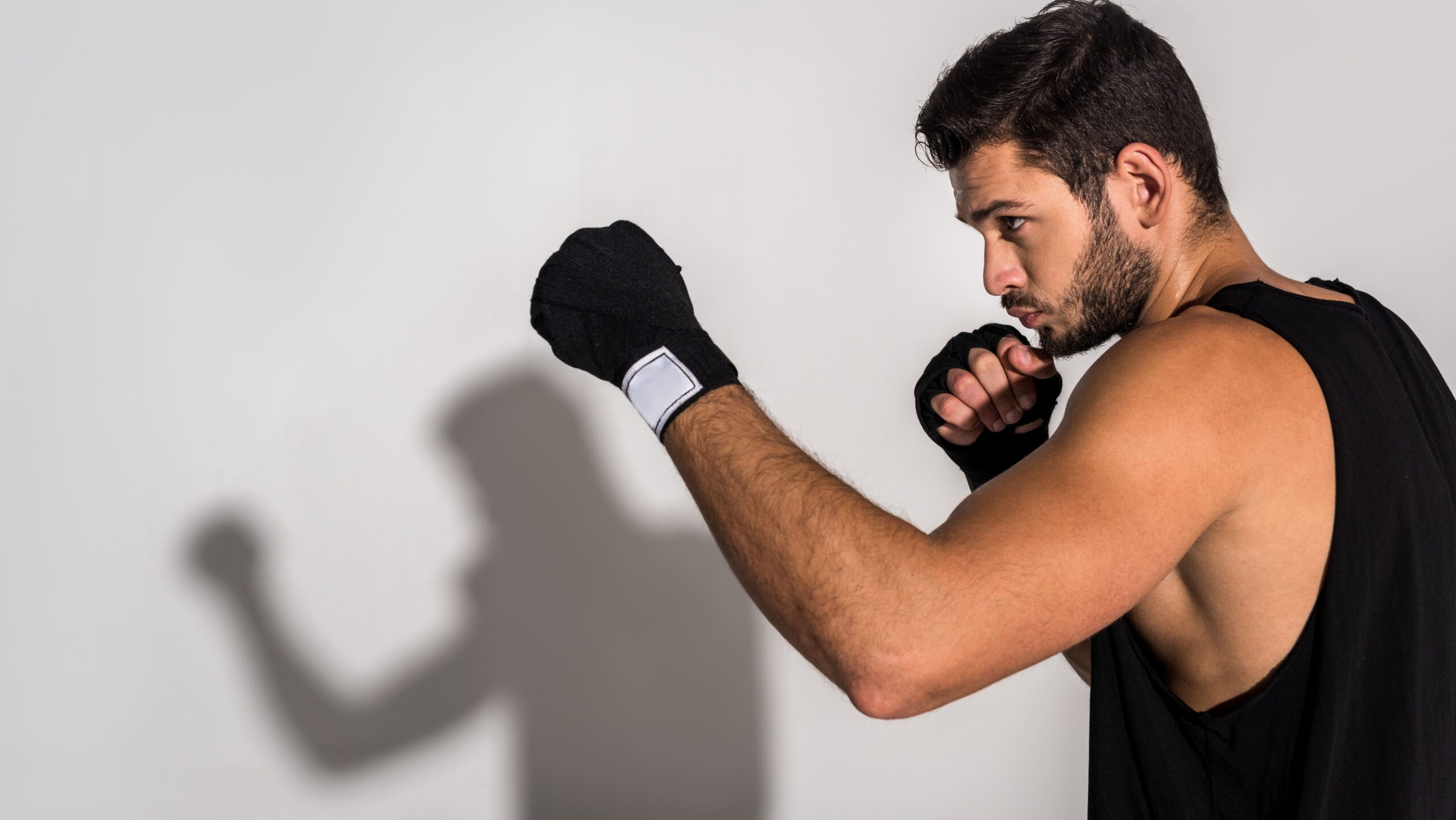 Homem com luva de boxe preta e faixa no punho em estúdio, em posição de ataque, com expressão focada, apontando o soco contra a frente em fundo claro.