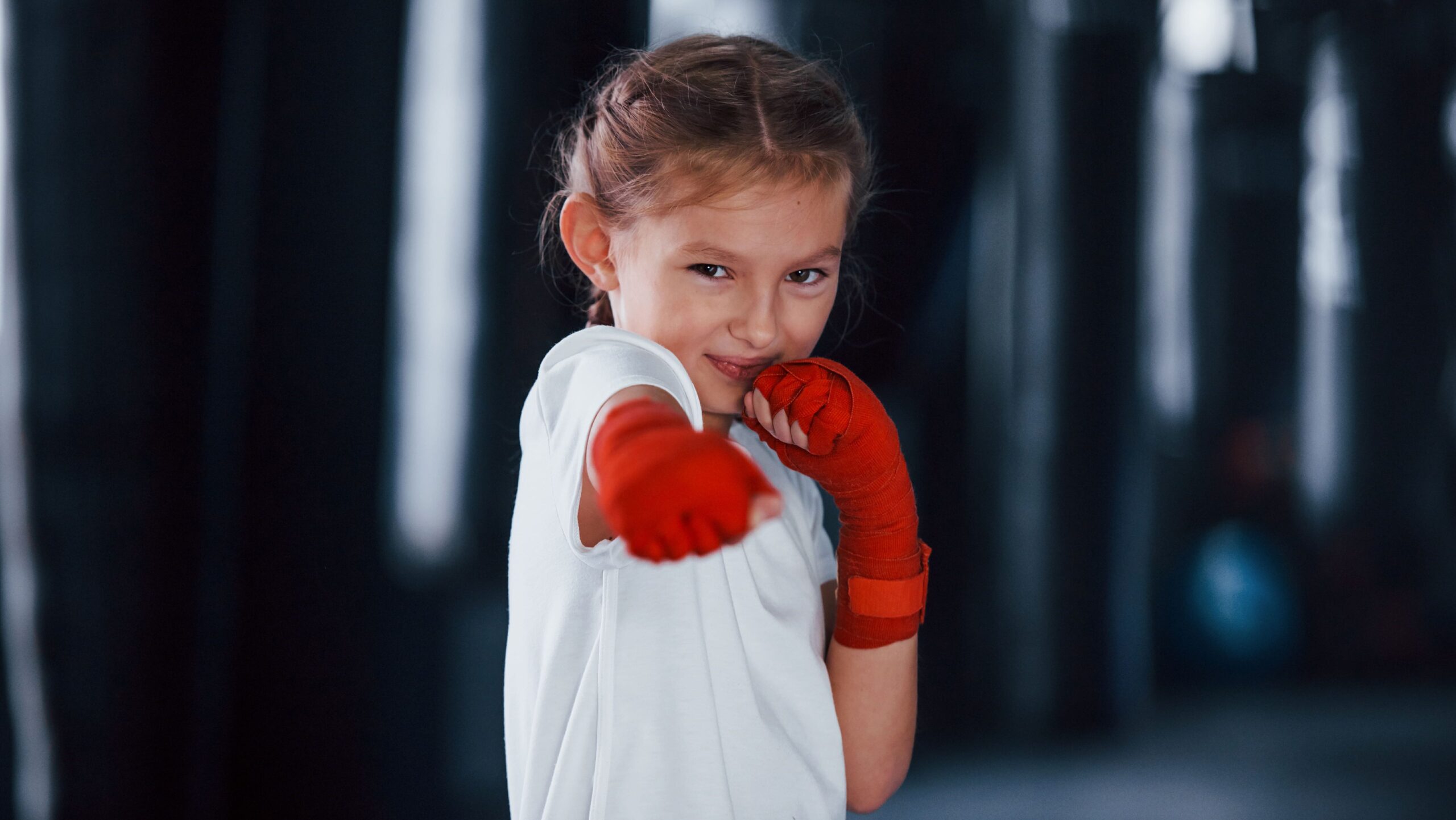 Menina com as mãos envolvidas em bandagens vermelhas simulando um soco em direção à câmera durante treino de combate.