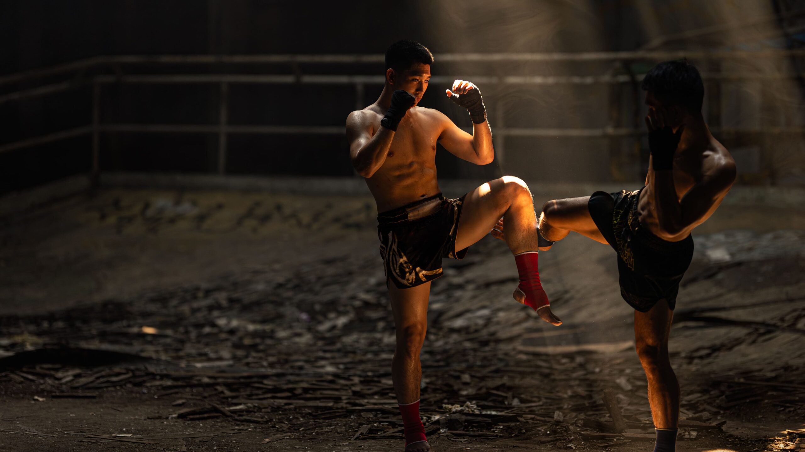 Dois atletas de Muay Thai treinando combate, com um executando um chute e o outro bloqueando o golpe com o joelho elevado.