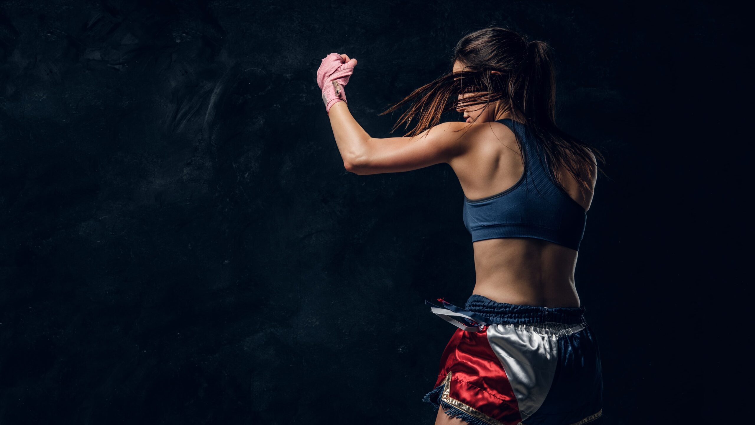 Mulher jovem treinando boxe em estúdio escuro, com luvas cor-de-rosa, postura lateral e abdômen à mostra, preparando um golpe contra o fundo escuro