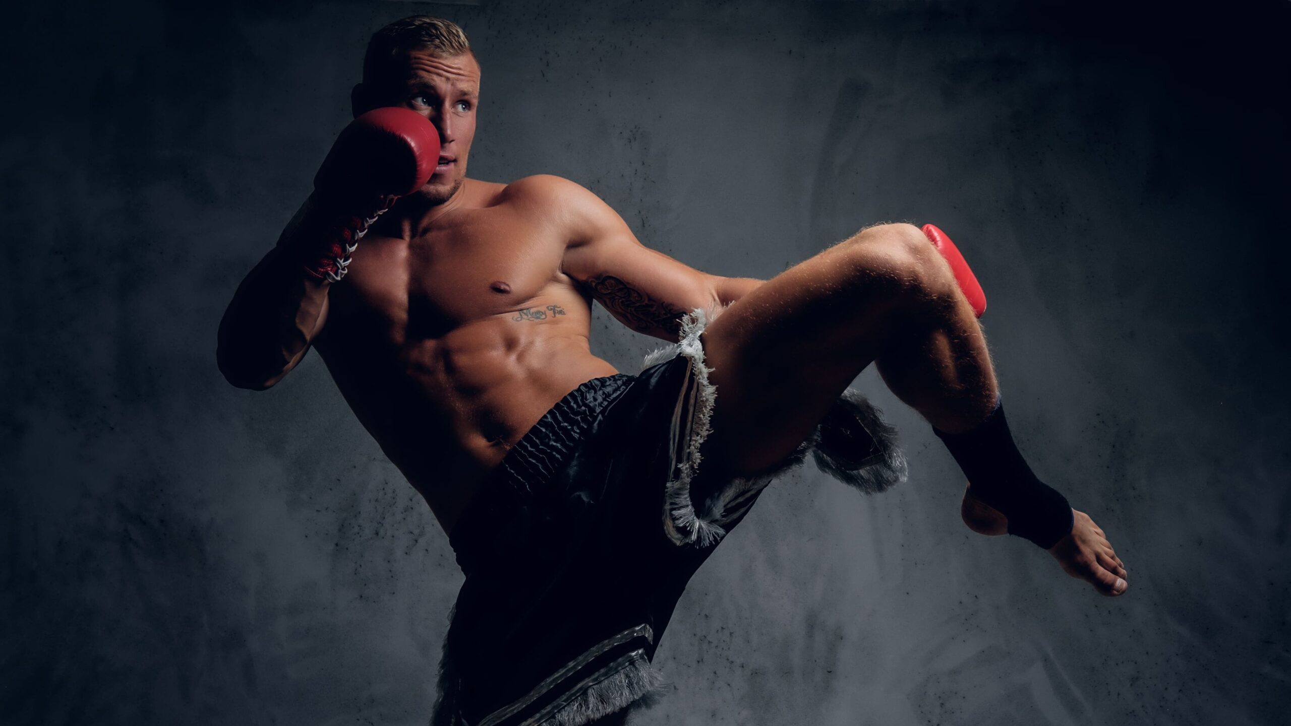 Homem praticando luta de boxe com luvas vermelhas, em posição de ataque, chutando ou preparando um golpe, em ambiente escuro com fumaça e clima intenso