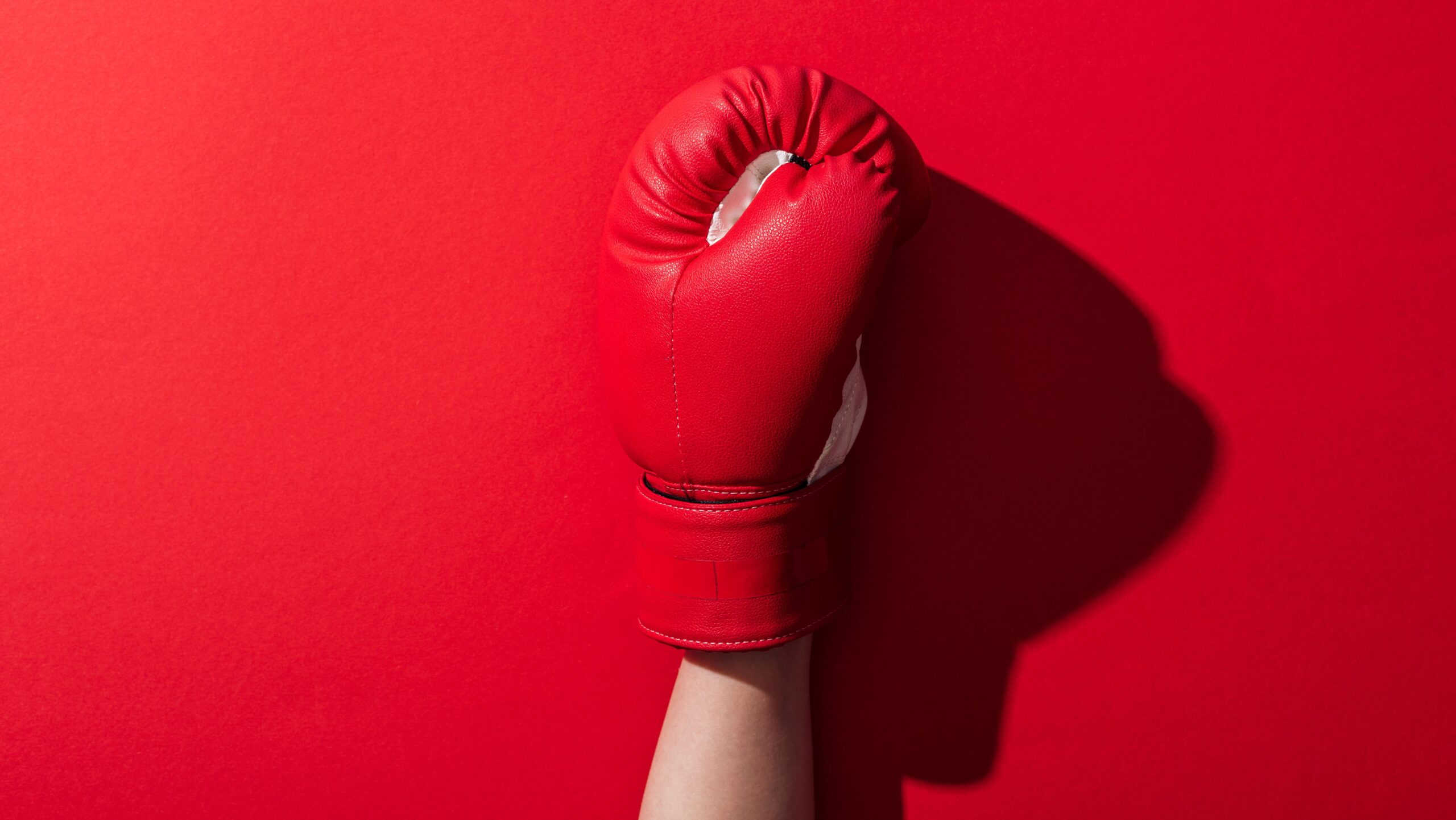 Mão com luva de boxe vermelha erguida em pose de combate, destacada contra fundo vermelho, com foco na textura do couro e no detalhe das costuras.