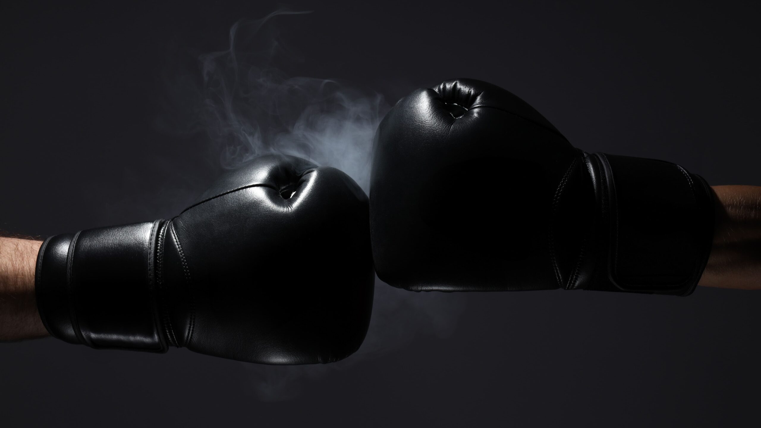 Duas luvas de boxe pretas se tocando no ar com uma leve névoa saindo do ponto de impacto contra fundo escuro.