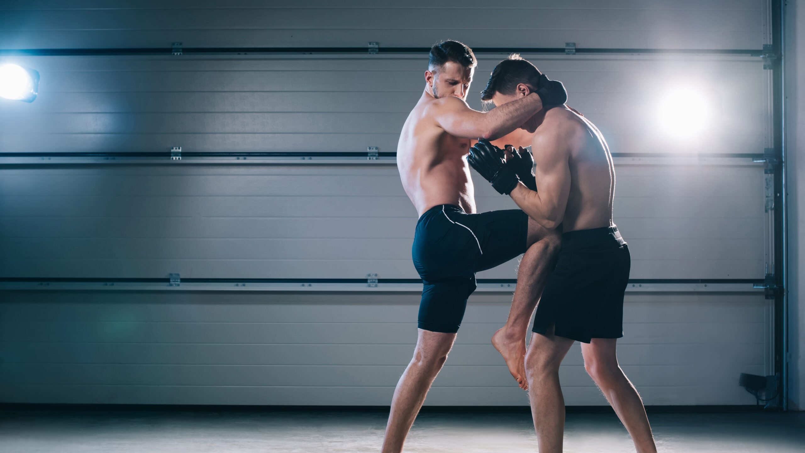 Dois atletas de muay thai sem camisa e descalços executando a luta agarrada. O atleta da esquerda segura a nuca do oponente para controlar sua postura enquanto eleva o joelho para aplicar uma joelhada reta no tronco, sob iluminação dramática de holofotes em estúdio de treino.