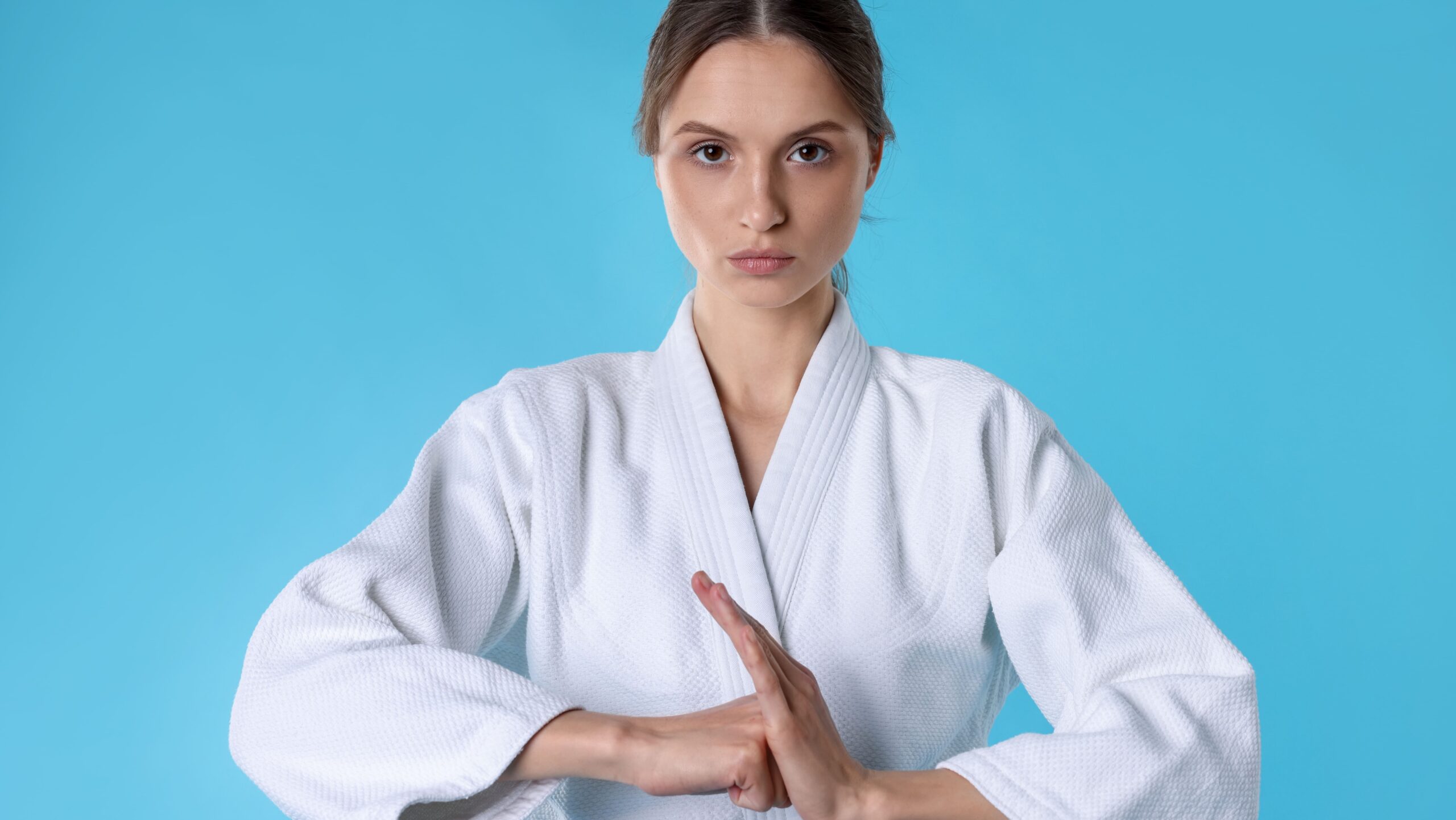 Mulher vestida com kimono branco faz sinal de “pare” com as mãos e expressão séria, em estúdio com fundo azul liso, pronto para comunicação visual