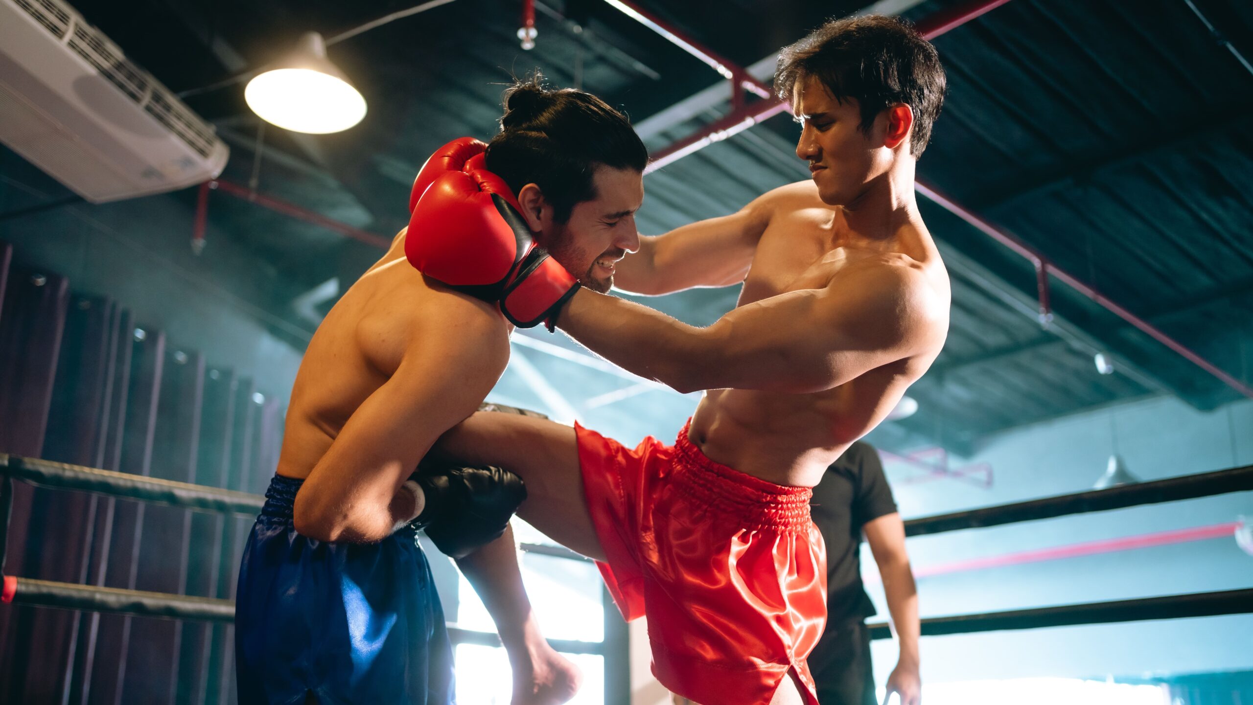 Dois lutadores de muay thai no ringue, onde o lutador de calção vermelho executa um duplo controle de nuca e aplica uma joelhada reta no oponente de calção azul.