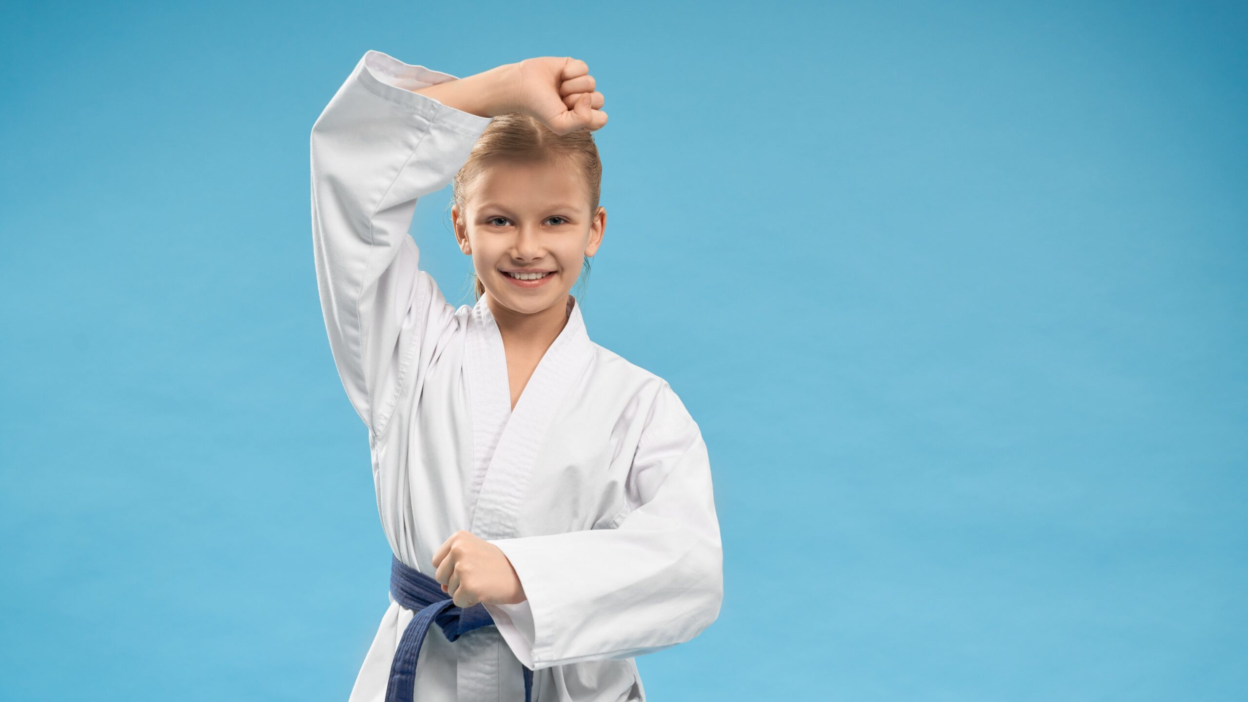 Menina sorridente vestindo quimono com faixa azul, executando movimento de defesa de cabeça contra fundo azul claro.