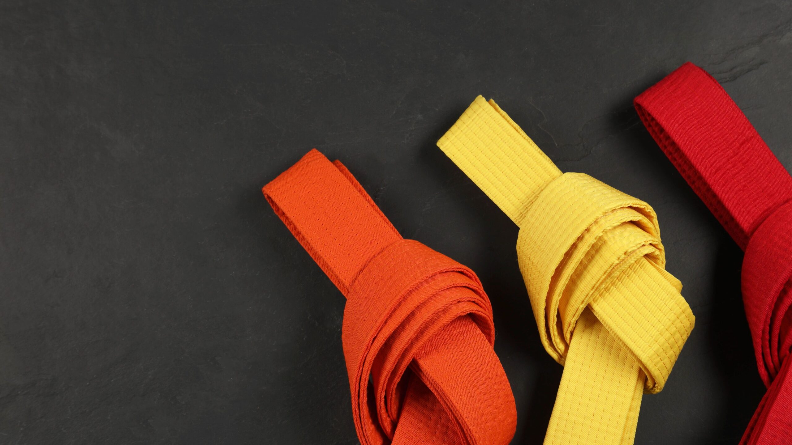 Três faixas dobradas de artes marciais nas cores laranja, amarela e vermelha organizadas sobre uma superfície escura.