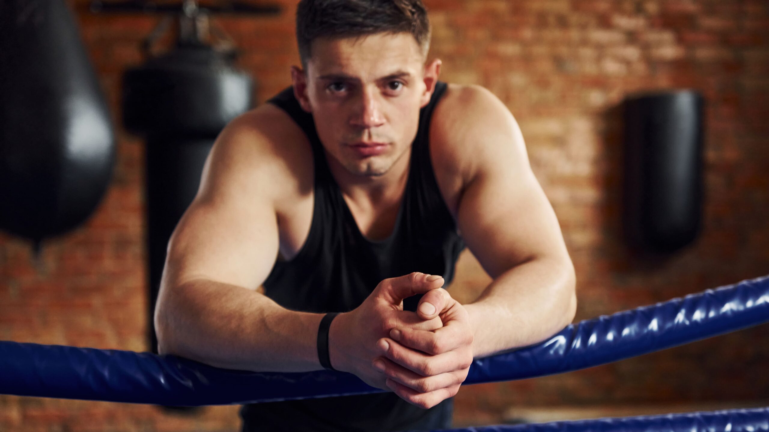 Homem jovem em treino de boxe, apoiado na grade azul do ringue, com postura firme e expressão focada, usando camiseta regata preta em ambiente com tijolos ao fundo.