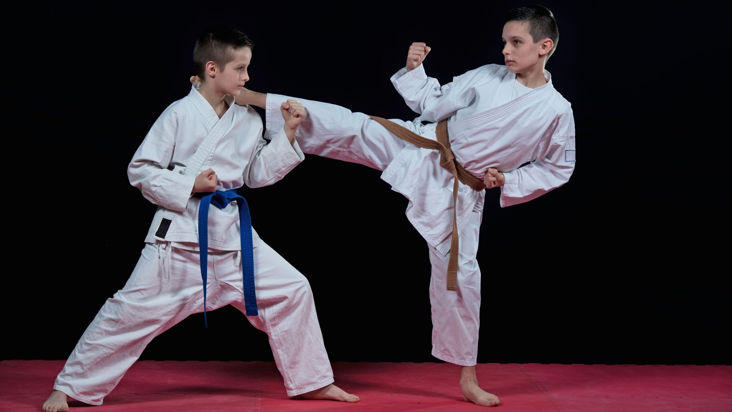 Dois meninos de quimono treinando no tatame vermelho, com um executando um chute lateral alto enquanto o outro mantém a guarda.