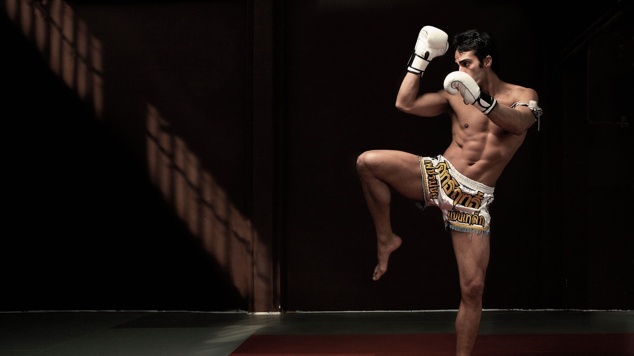 Atleta em academia de treino executando movimento de guarda com joelho elevado, usando luvas brancas e shorts tradicionais de Muay Thai.