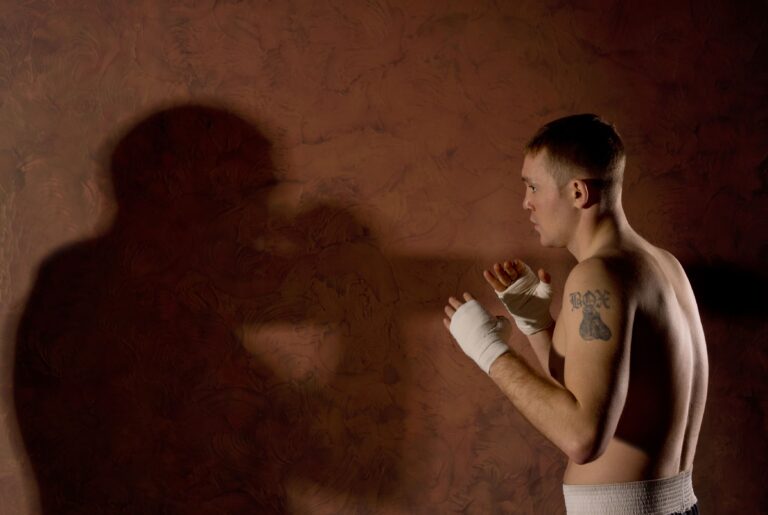 Homem sem camisa com curativos nos punhos, tatuagem no peito e expressão focada, em um cenário com fundo marrom e iluminação dramática, sugerindo treino de luta ou preparação para boxe.