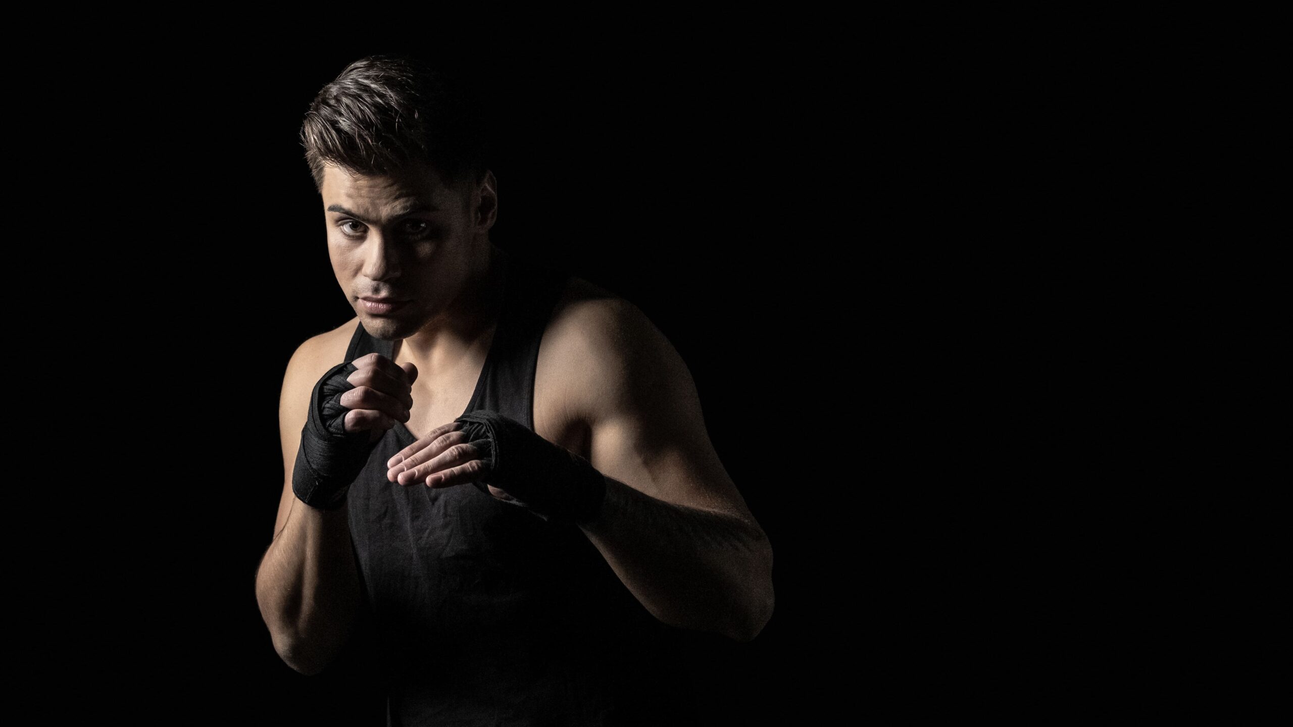 Homem em estúdio com roupa escura, segurando luvas de boxe e em posição de combate, com iluminação dramática contra fundo preto, pronto para lutar.