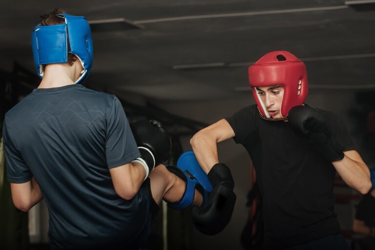 Dois lutadores em um ringue de boxe, usando luvas e capacetes azul e vermelho, treinando golpes e defesas durante uma sessão de treino em um tatame/oficina esportiva.