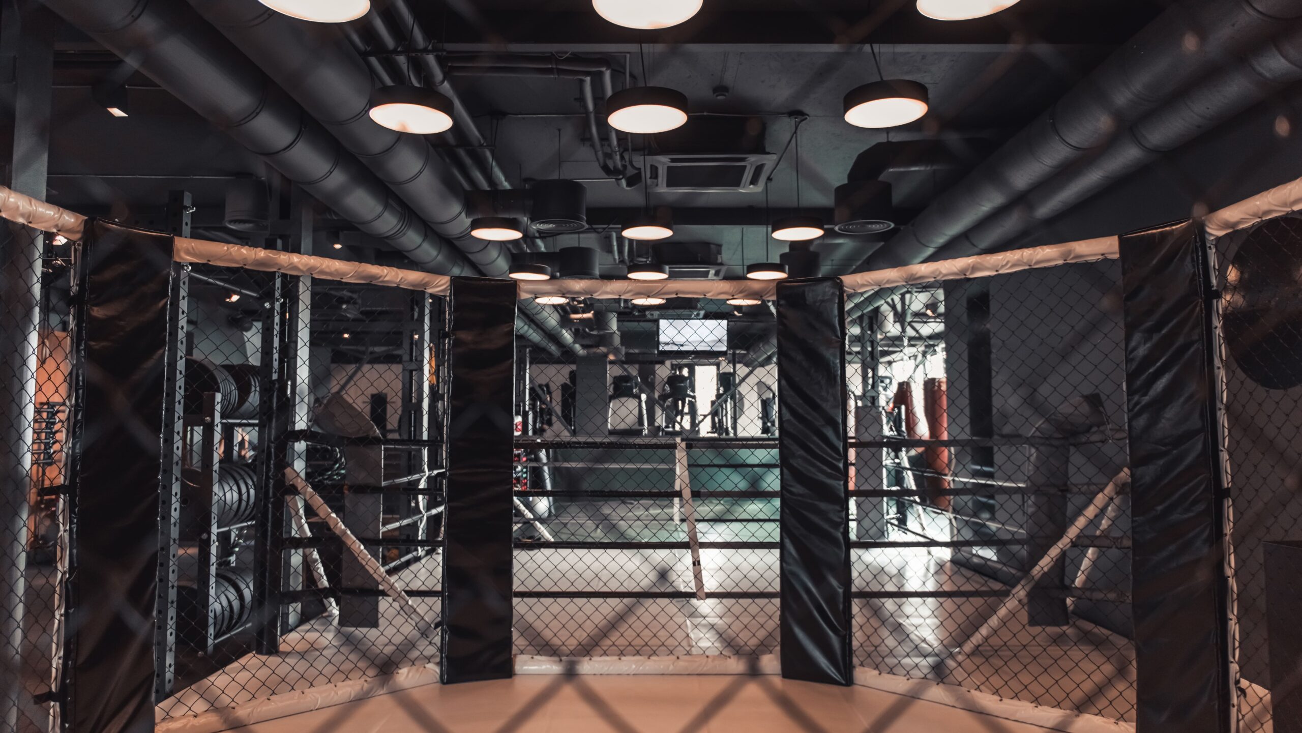 Interior de uma sala de treino ou luta com boxe de MMA, grades e duas portas abertas, iluminação do teto e piso de borracha em ambiente industrial