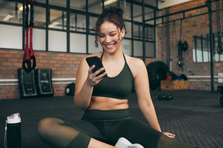 Mulher sorrindo enquanto está sentada no chão de uma academia de treino e utiliza o celular.