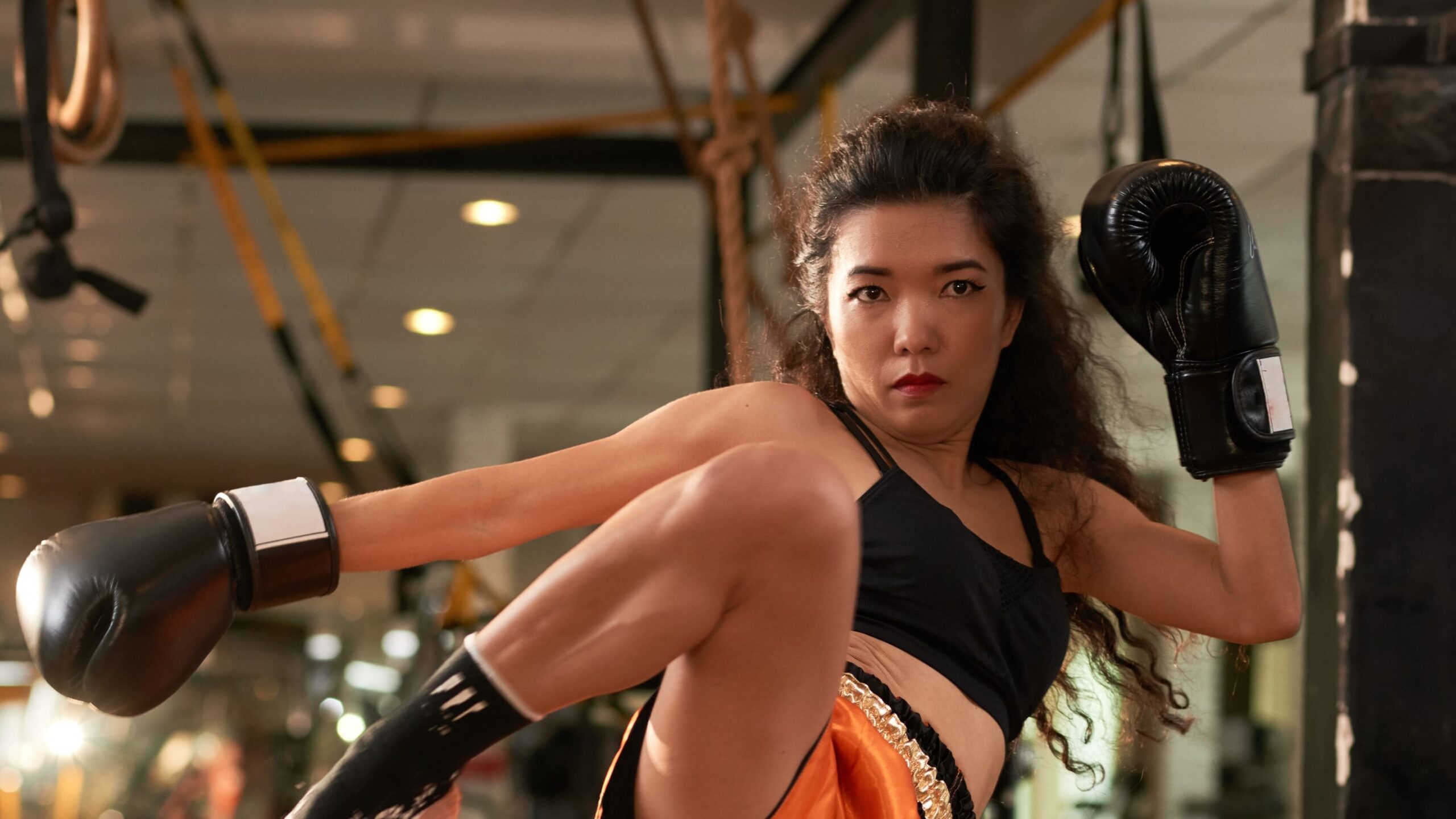 Mulher lutadora em academia de muay thai, usando luvas e halter, fazendo exercício de chute na frente de um saco e equipamentos no tatame