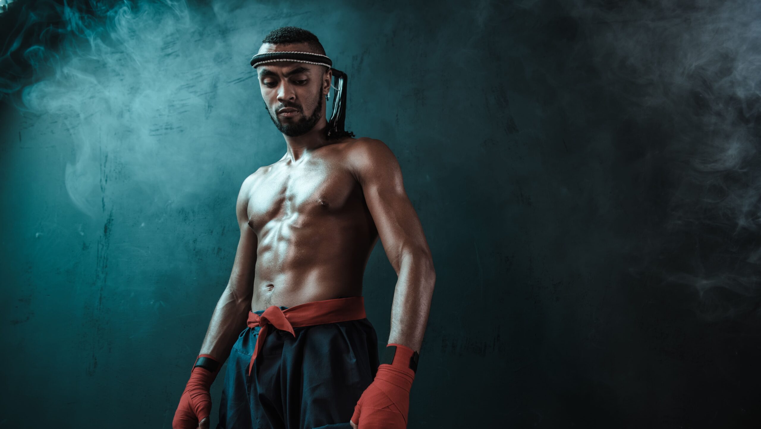 Homem atleta musculoso com faixa na cabeça e luvas de treino, usando shorts e postura de combate, em ambiente com fumaça e luz azul, estilo treino ou luta.
