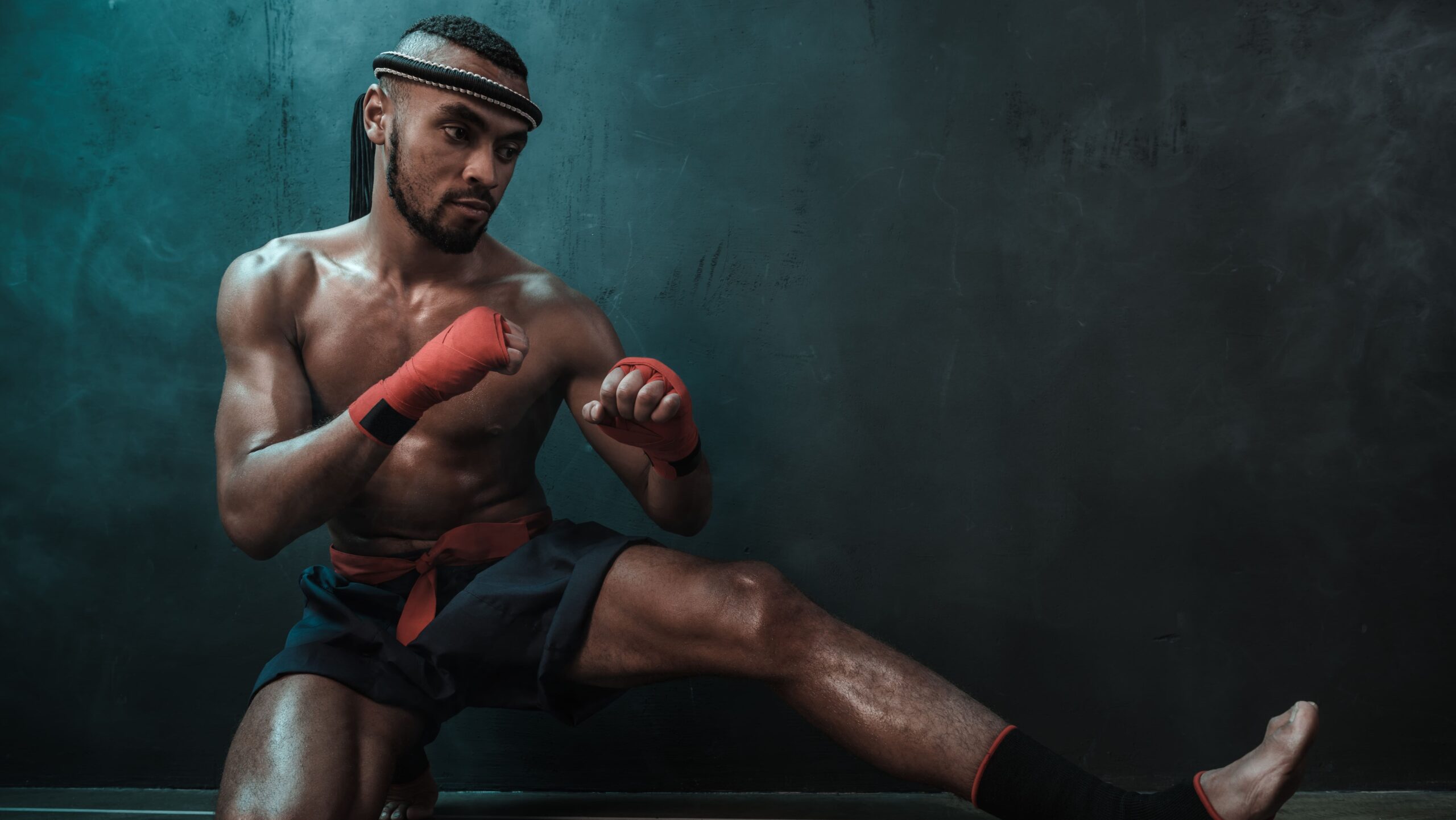 Homem atleta de muay thai treinando em estúdio, em posição de combate ajoelhado e segurando luvas vermelhas com munhequeiras, com postura focada e fundo escuro e azul.