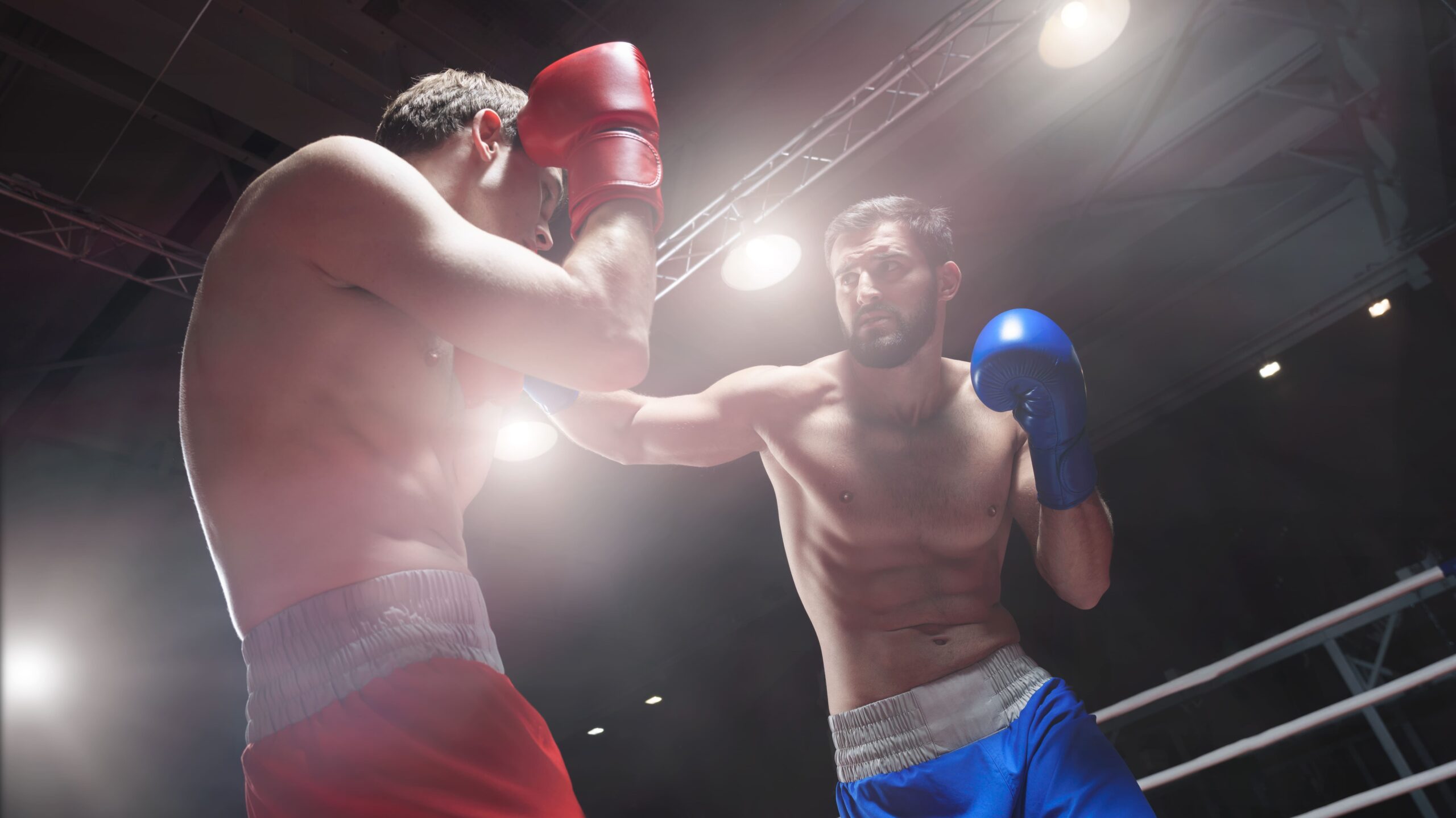 Luta de boxe no ringue: um boxeador com luvas vermelhas dá um golpe no oponente, que responde com luva azul, sob luzes fortes do ginásio