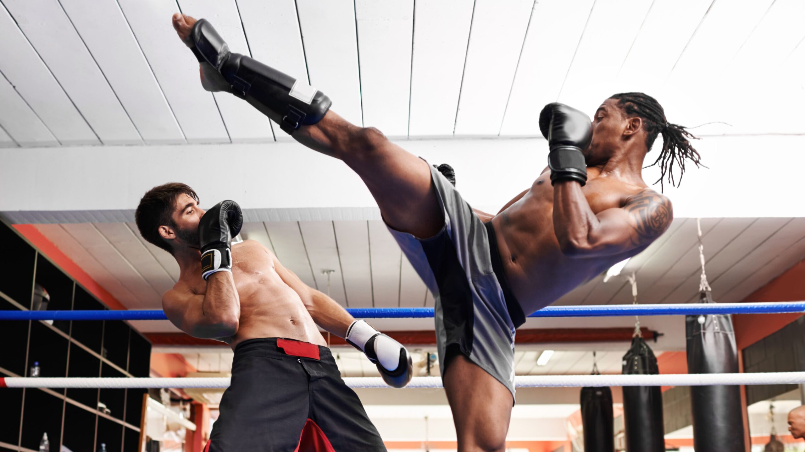 Lutadores de boxe em um ringue, com um atleta usando bandagens e luvas enquanto dá um golpe alto com a perna, no ambiente de treino
