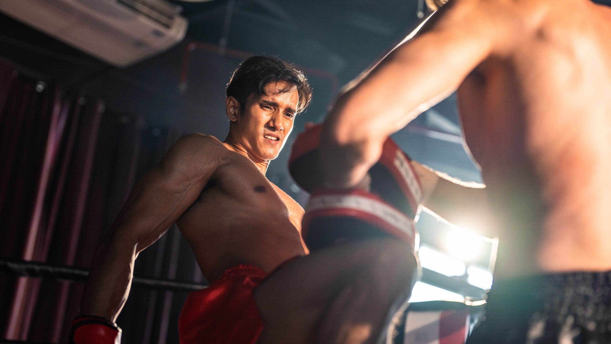 Lutador de muay thai em um ringue de luta, com luzes dramáticas no ginásio, socando e mirando o adversário em primeiro plano, clima de competição e intensidade