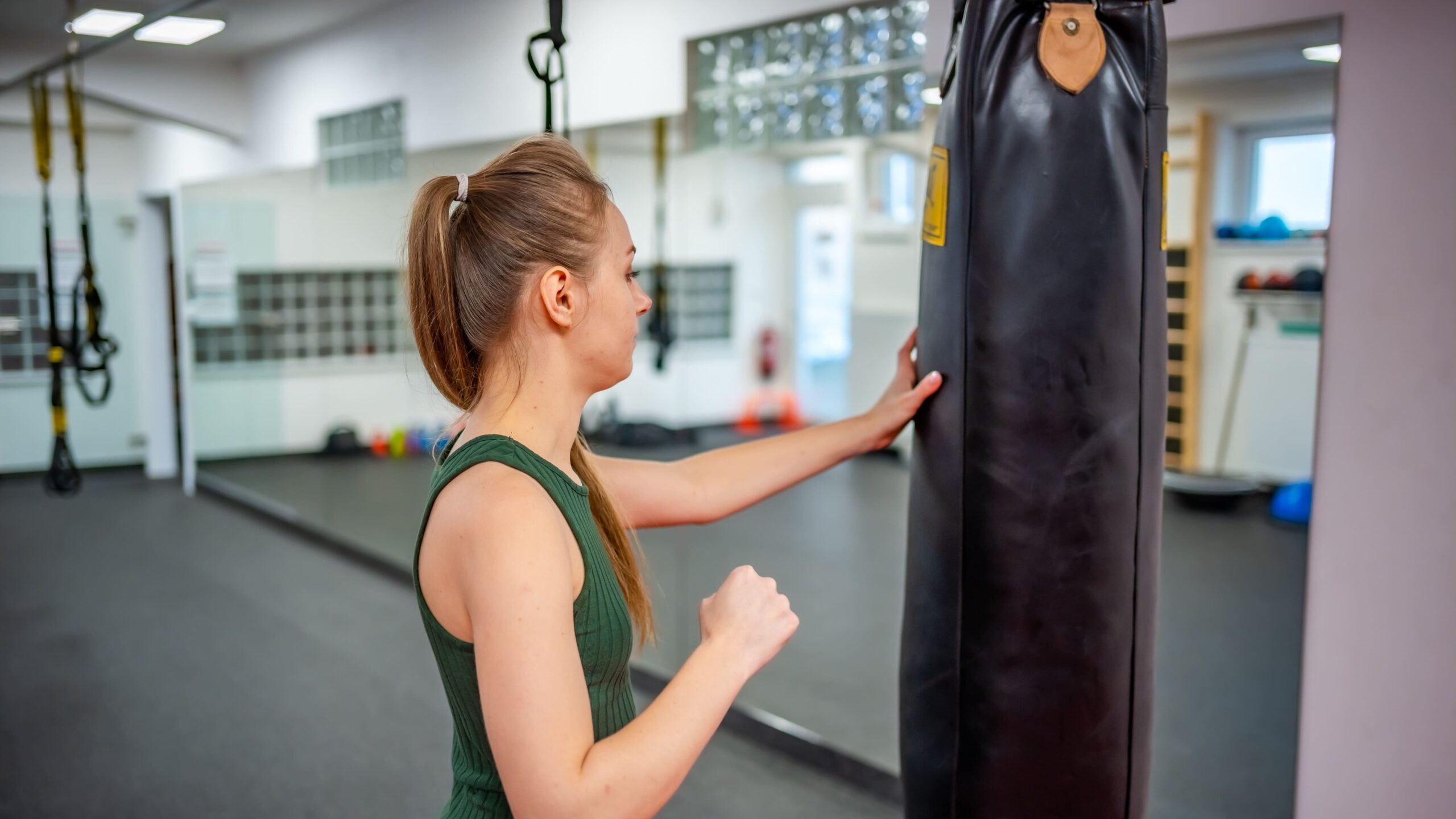 Mulher em uma sala de treino segura um saco de pancadas suspenso no boxe, em posição lateral, com postura de preparação para o exercício