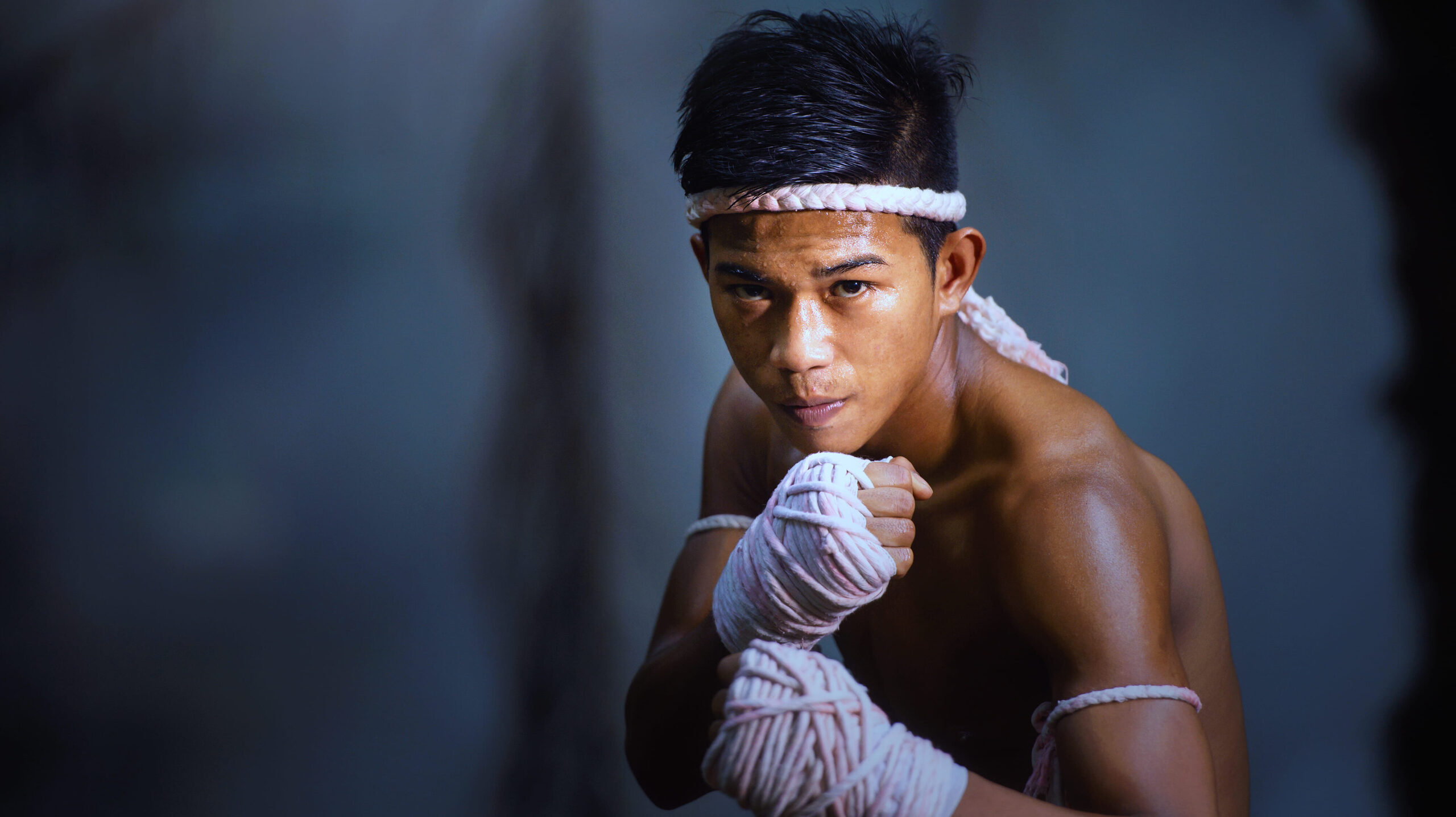 Lutador de boxe com faixa branca na testa e bandagens nas mãos, em pose de treino no ringue, com expressão focada e fundo desfocado azul. Foto em estilo esportivo e dramático.