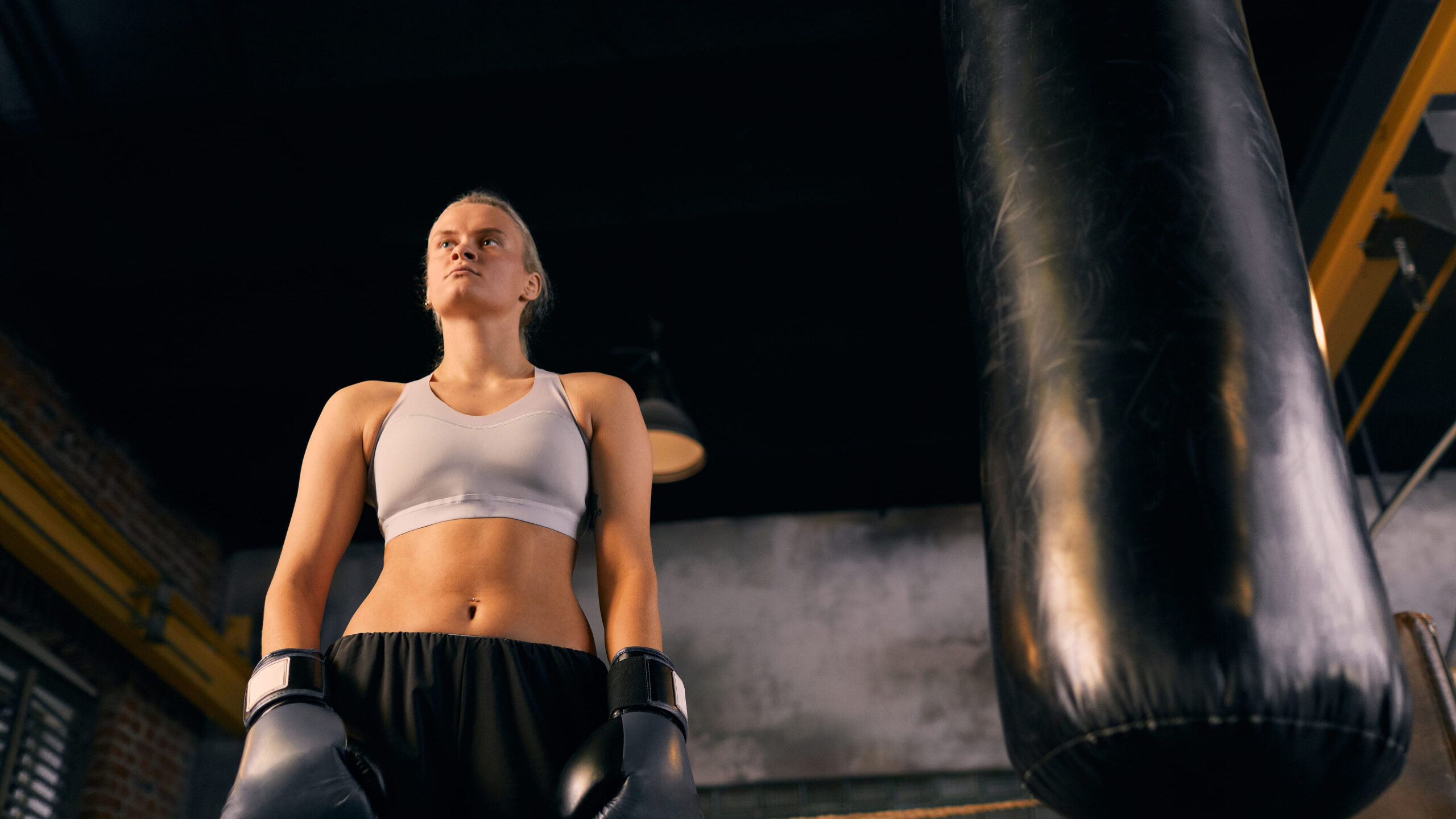 Mulher atleta em treino de boxe em academia, usando luvas de boxe pretas e top esportivo branco, posicionada ao lado de um saco de pancadas com iluminação de treino.