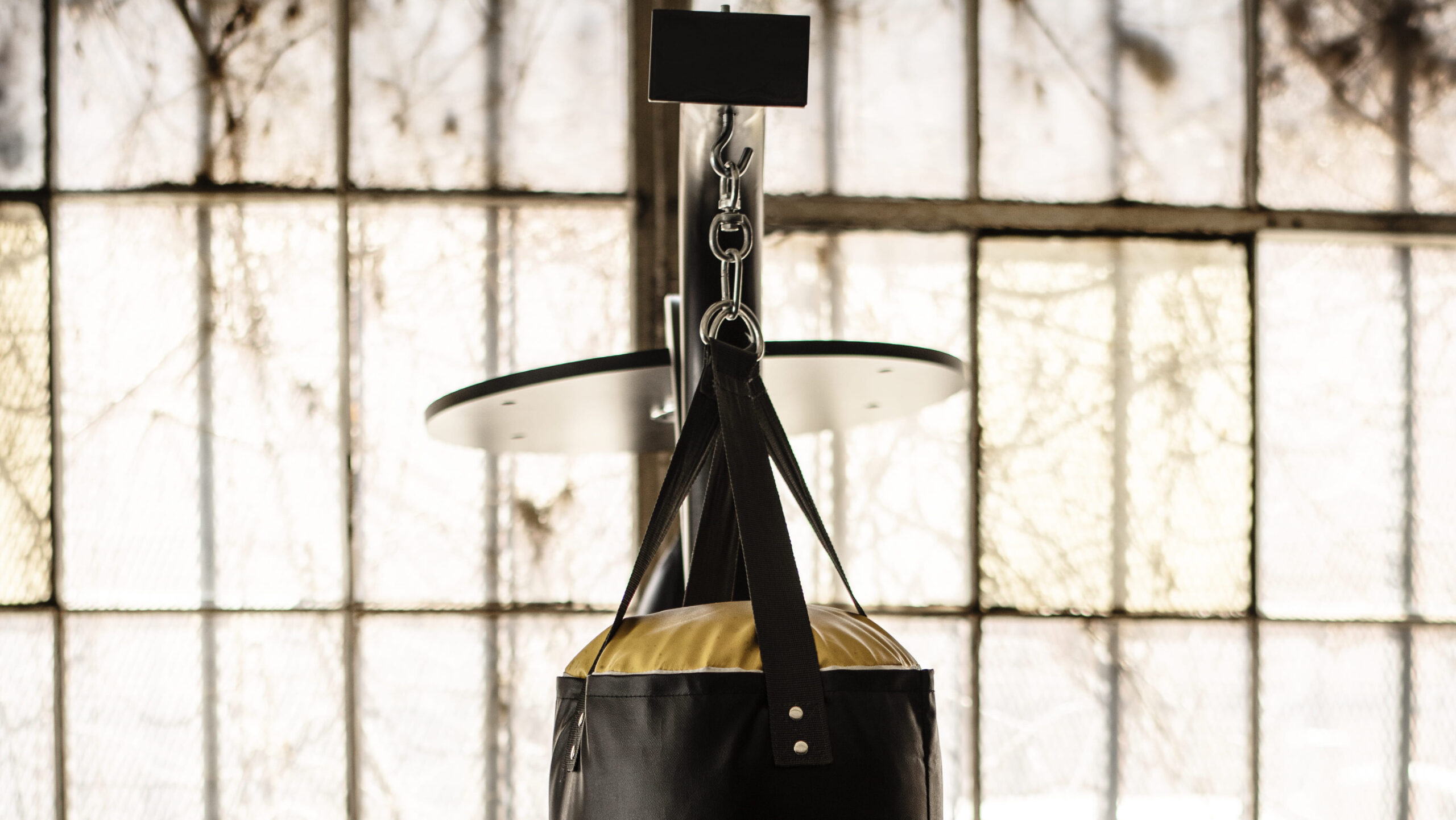 Saco de pancada de boxe preto com alça e suporte de metal, preso ao teto, em ambiente com parede de tijolos e luz natural.