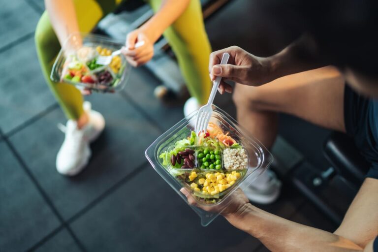 Pessoas separando e montando uma salada em pote de plástico com legumes coloridos como ervilha, milho, folhas e cenoura, em ambiente externo.