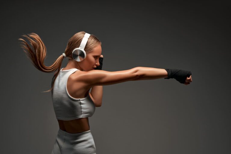 Mulher com roupa esportiva e fones de ouvido, praticando treino de boxe com os punhos erguidos em estúdio, usando luvas