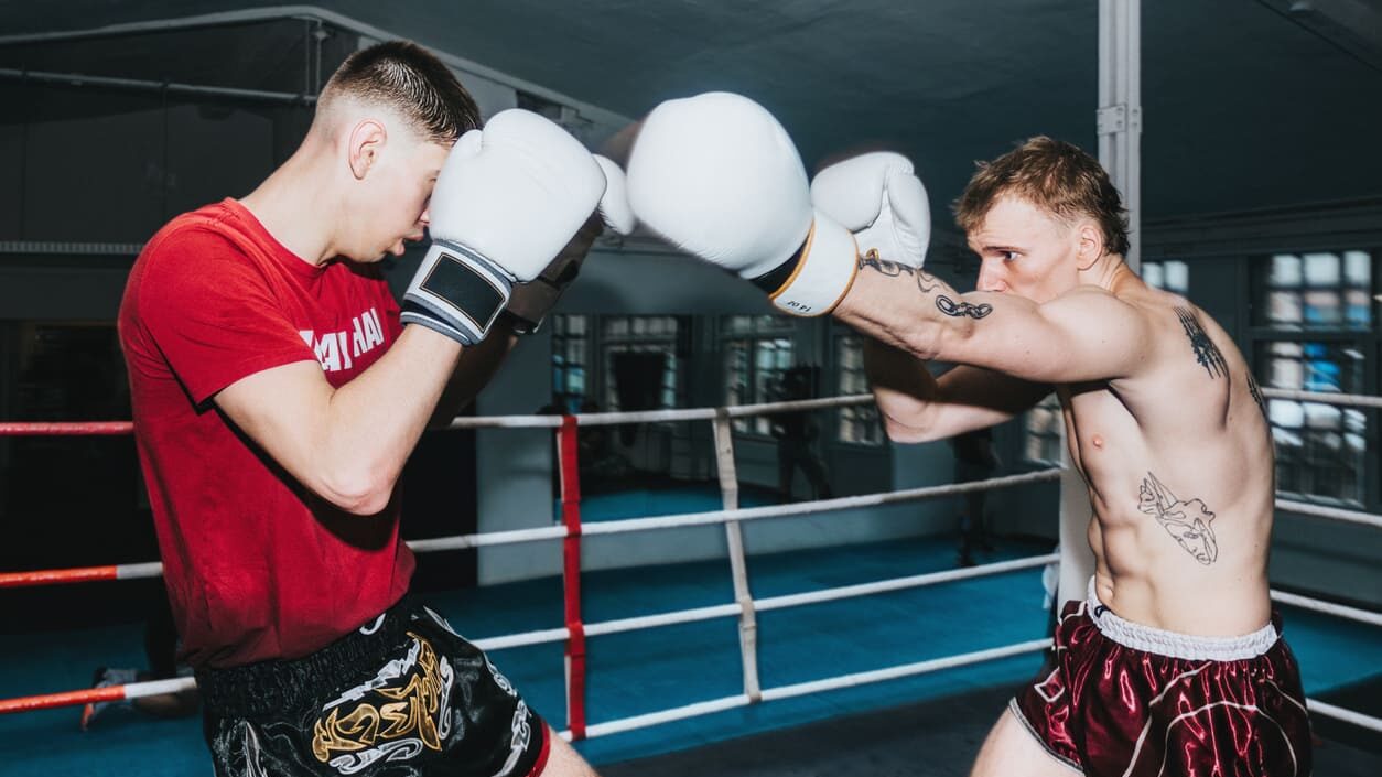 Dois lutadores de muay thai em um ringue com luvas de treino, um em posição defensiva e o outro preparando um golpe, em cena de combate.