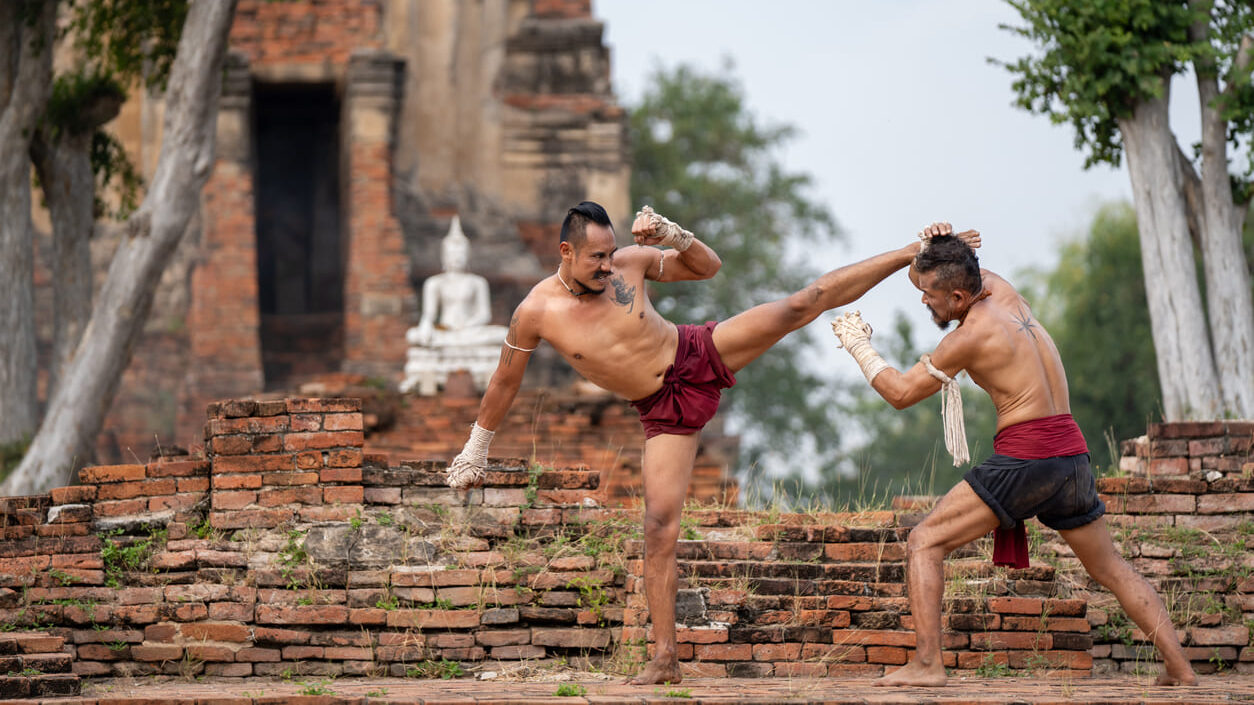 Dois lutadores tradicionais se enfrentam em uma disputa de muay thai ao ar livre, com roupas típicas e postura de combate, diante de ruínas de pedra e árvores.