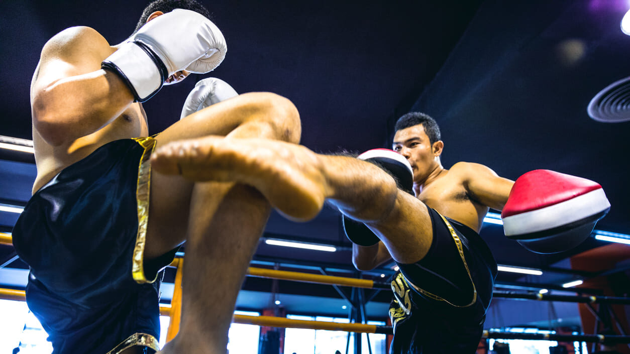 Dois lutadores de muay thai em um ringue, com um golpe sendo desferido no corpo, luvas brancas e botas esportivas, em meio à torcida e luzes do ginásio